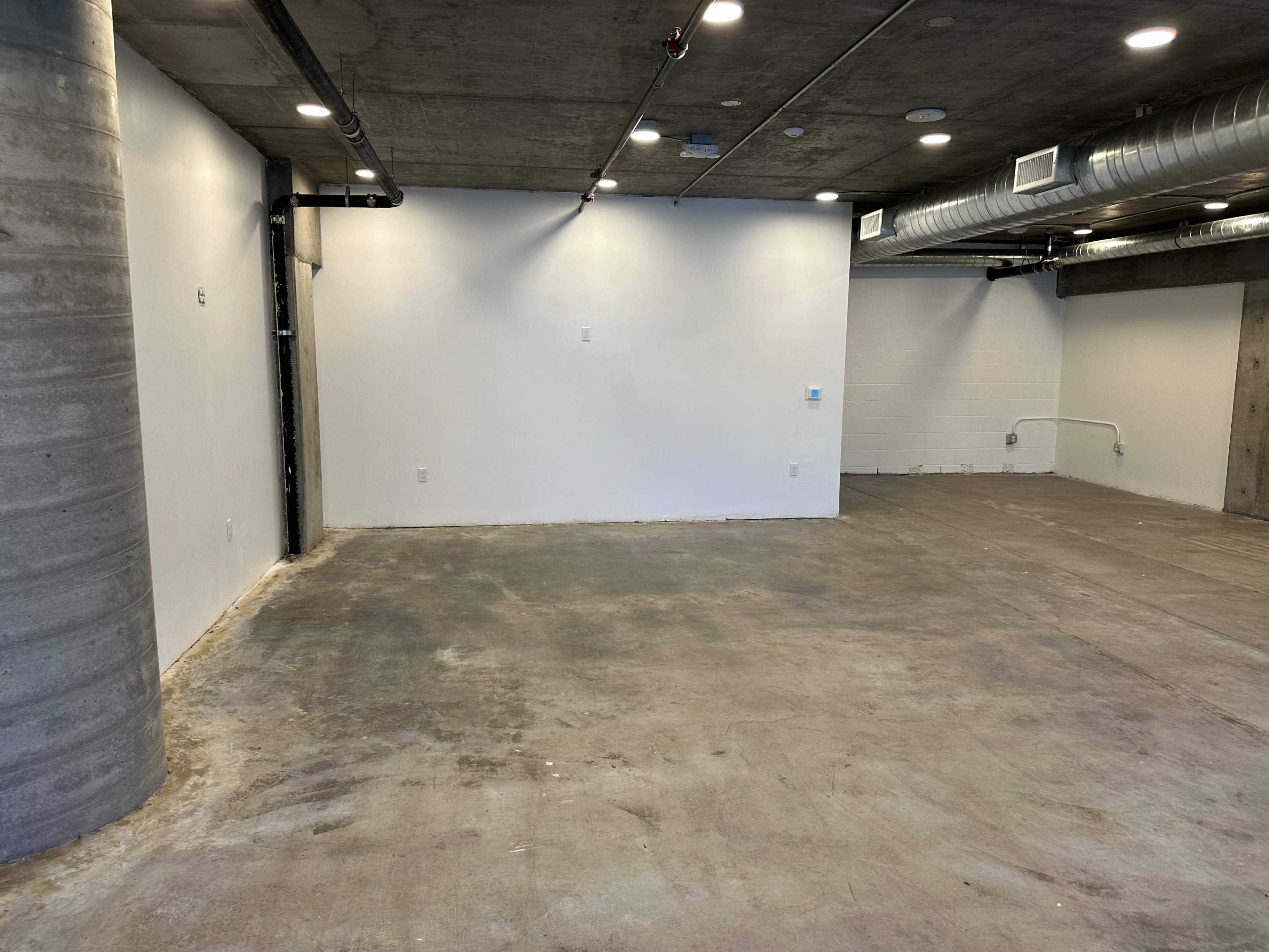 The image shows an empty interior space with concrete floors and white walls, featuring exposed pipes and a pillar on one side.