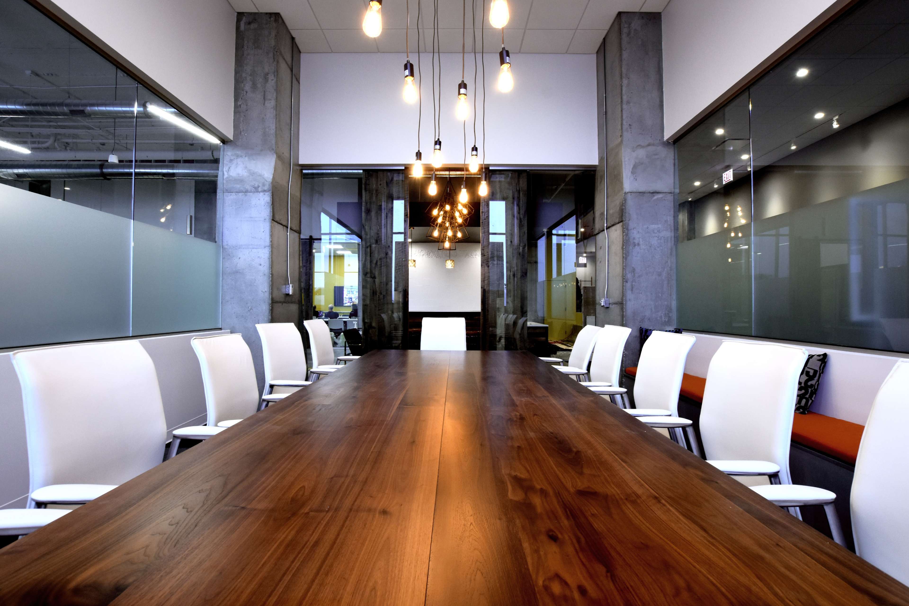 A long wooden conference table with white chairs is illuminated by pendant lights in a modern meeting room.