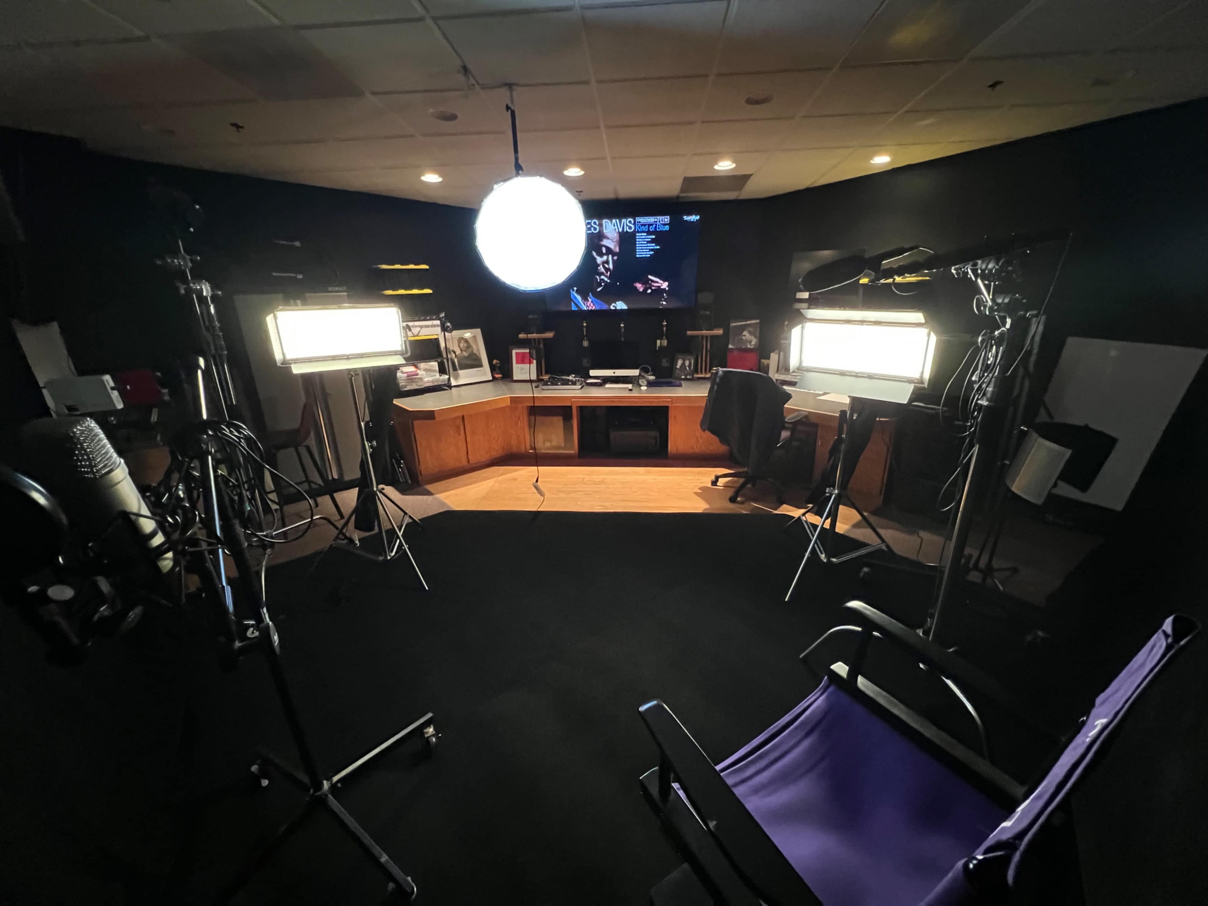 A video recording studio with lighting equipment, a desk, and a large screen in the background.