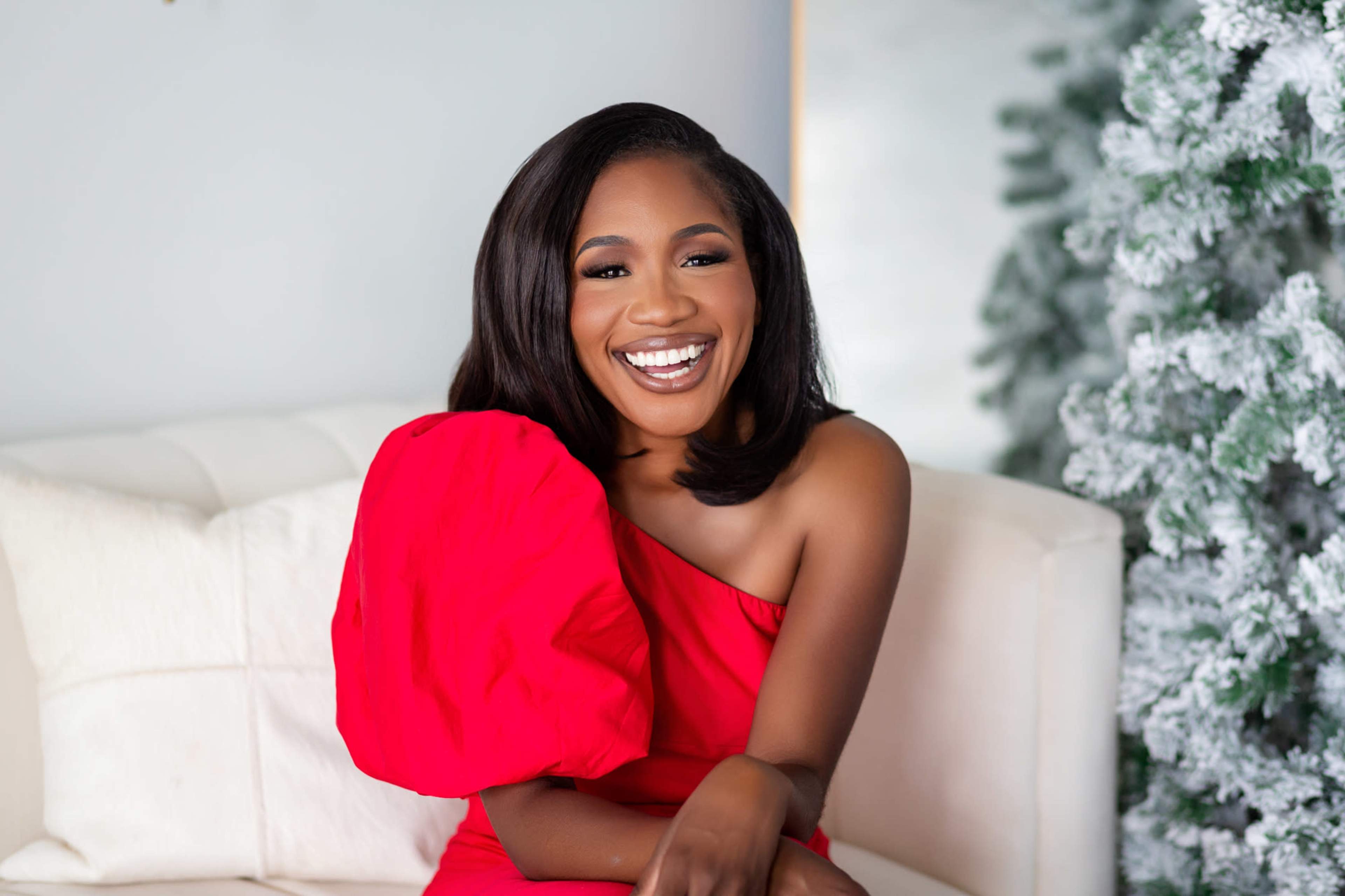 A woman in a red dress sits on a white couch, smiling in front of a decorated Christmas tree.