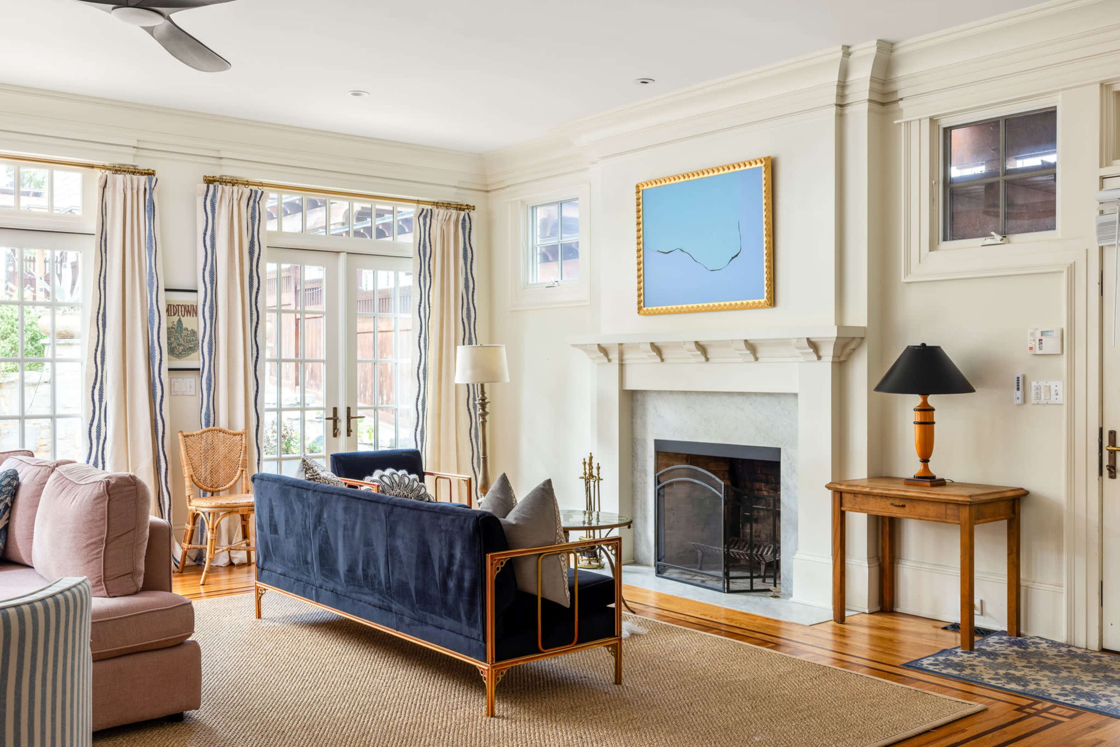 The image shows a bright living room featuring a blue sofa, a light-colored rug, a fireplace with a mantle, and large windows that let in natural light.