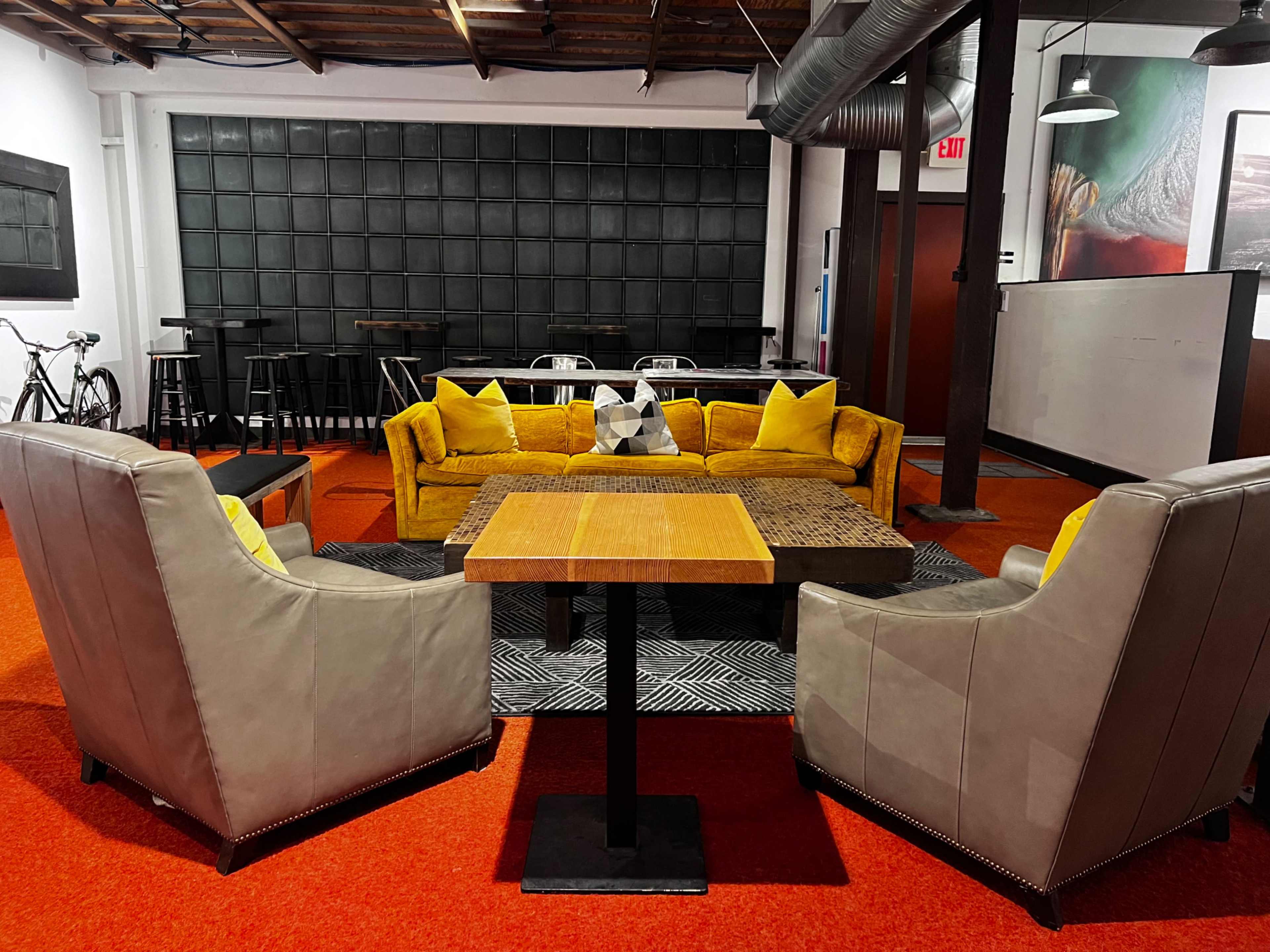 A modern lounge area features a yellow couch and gray armchairs arranged around a wooden coffee table on red carpet flooring.