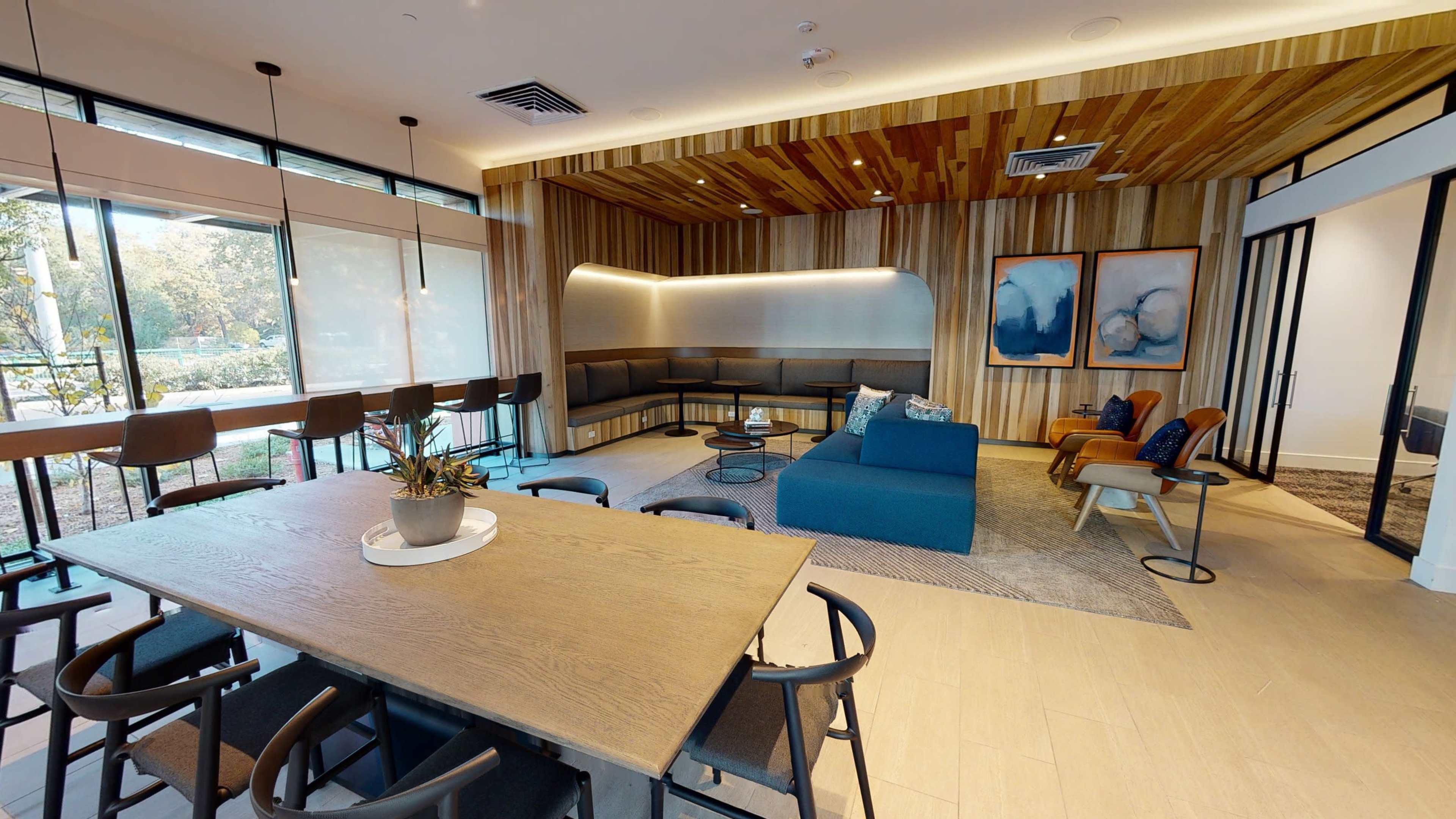 A modern lounge area with a large wooden dining table, a blue sectional sofa, and a cozy seating arrangement against a wood-paneled wall.