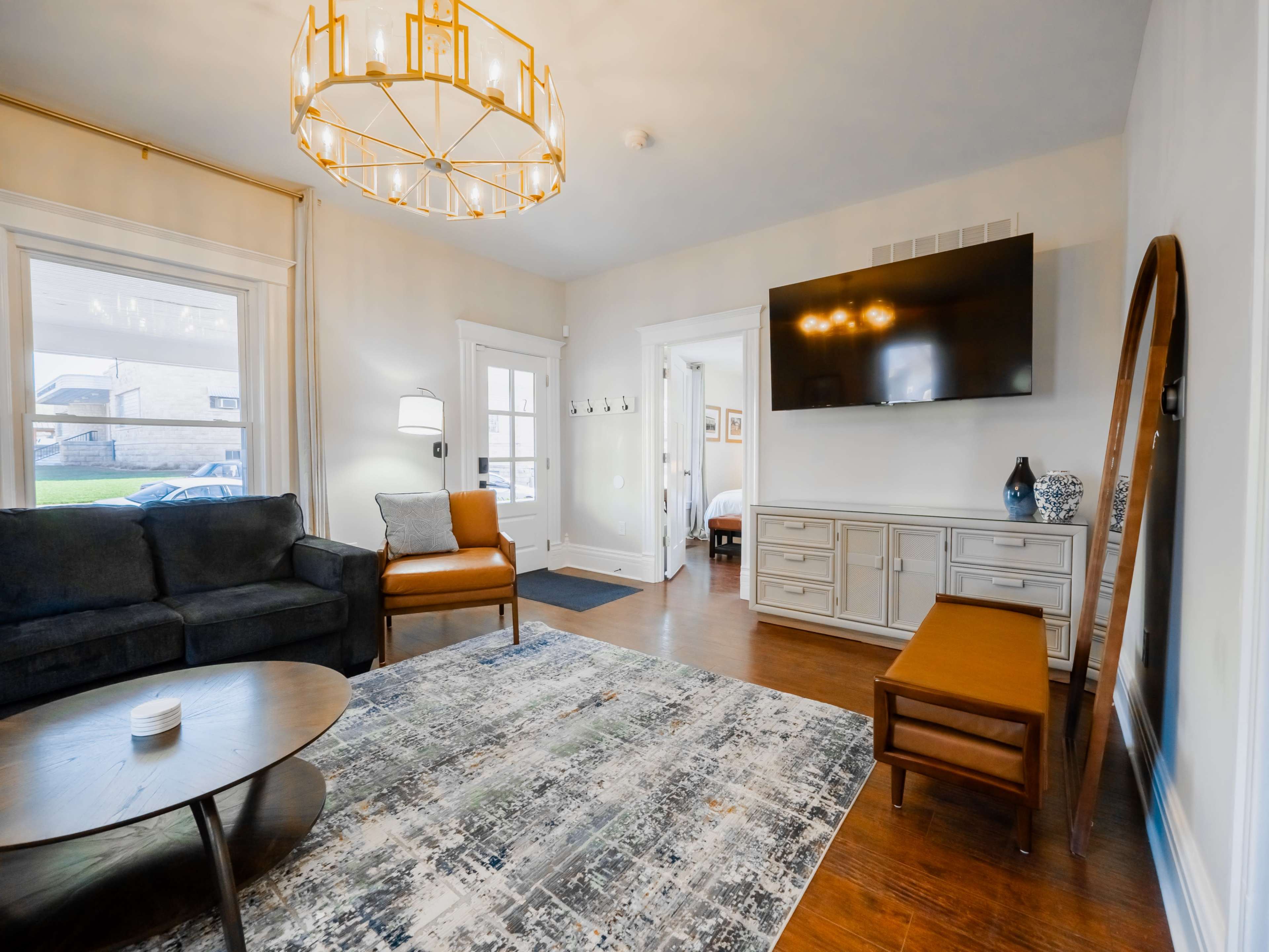 The image shows a modern living room featuring a round coffee table, an armchair, a large wall-mounted TV, and a patterned rug on a wooden floor.