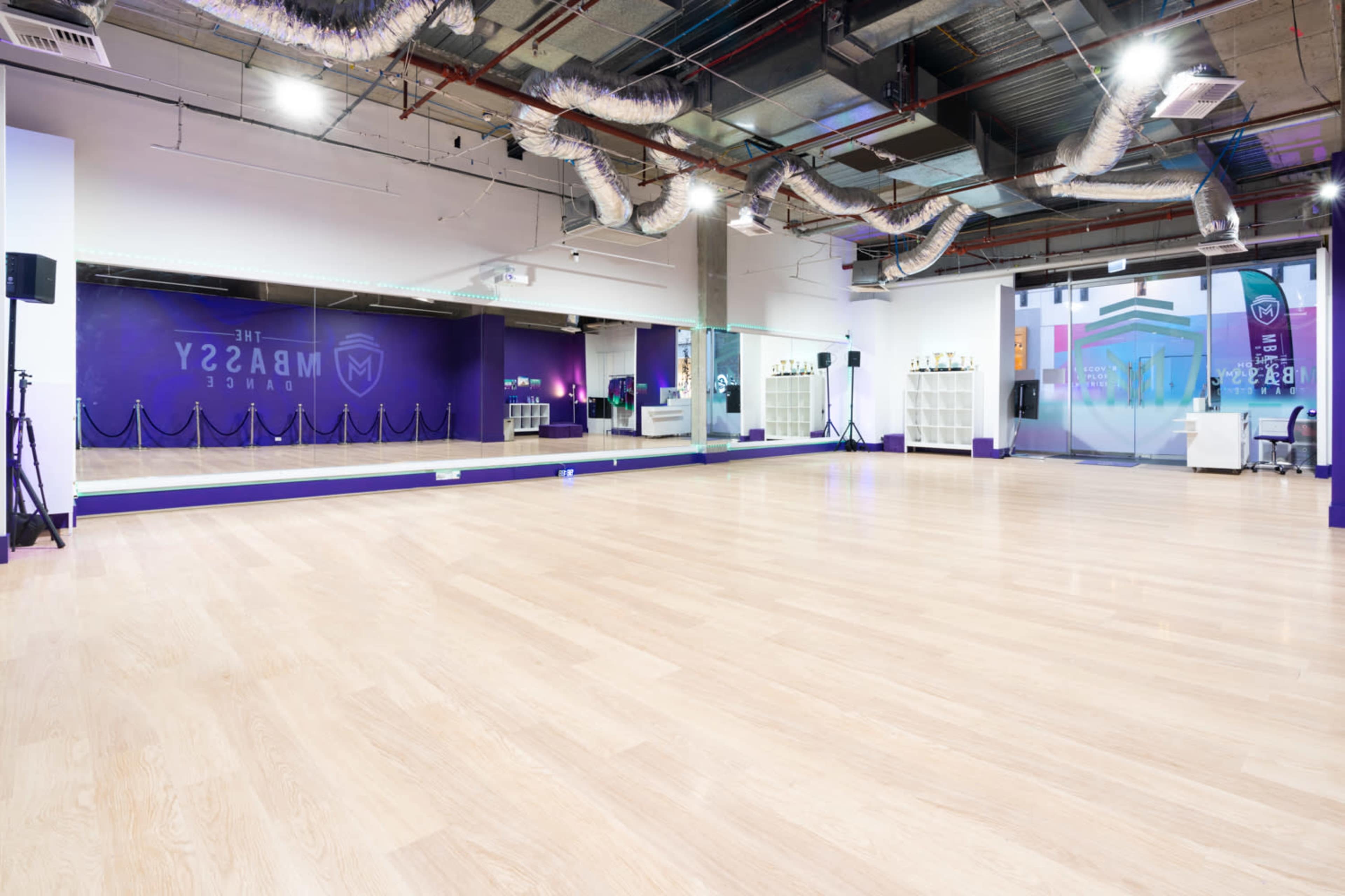The image shows an empty dance studio with wooden flooring, mirrored walls, and purple accents.