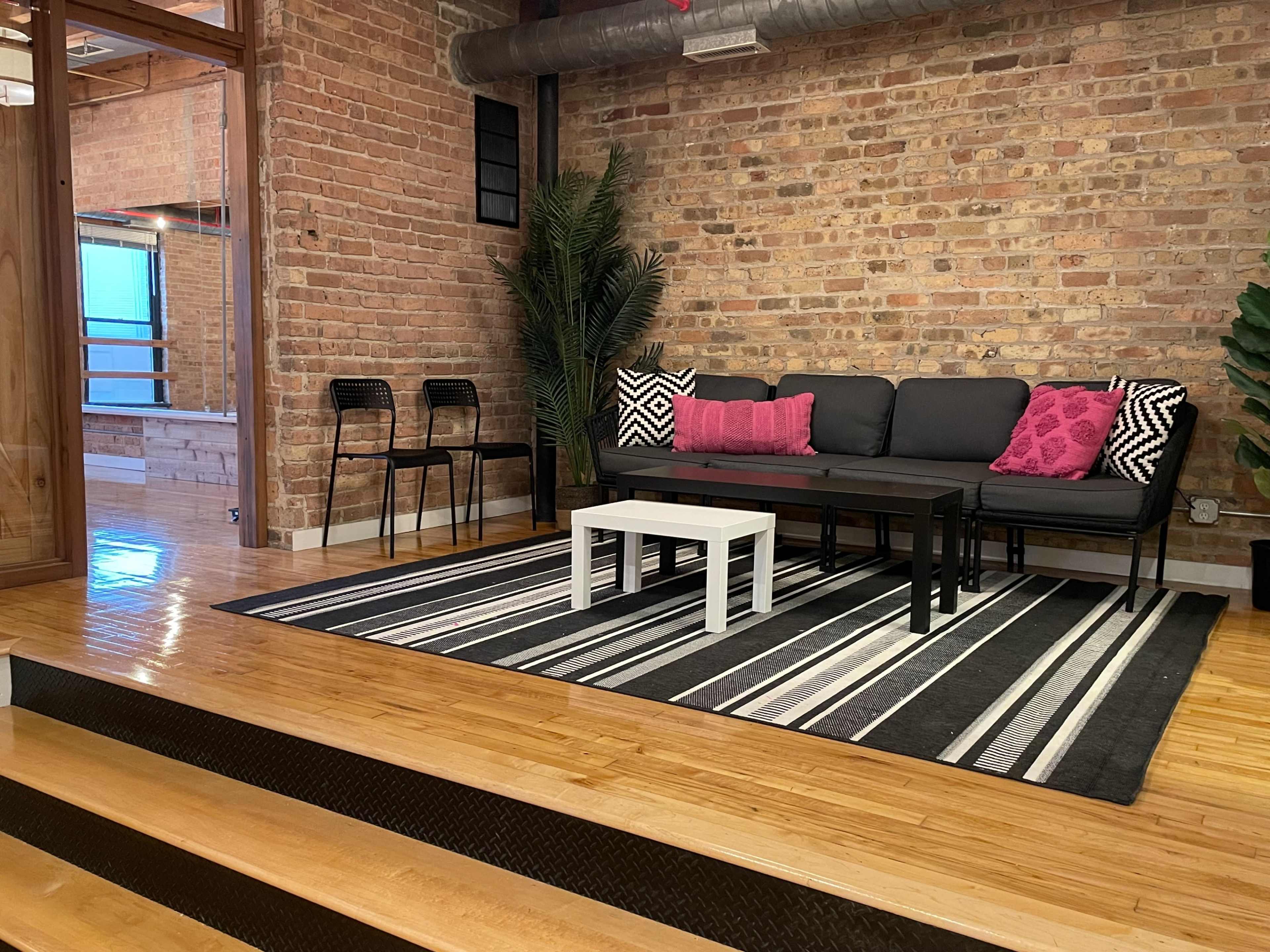 A modern living area features a black sofa with pink pillows, a striped rug, a white coffee table, and a backdrop of exposed brick walls.