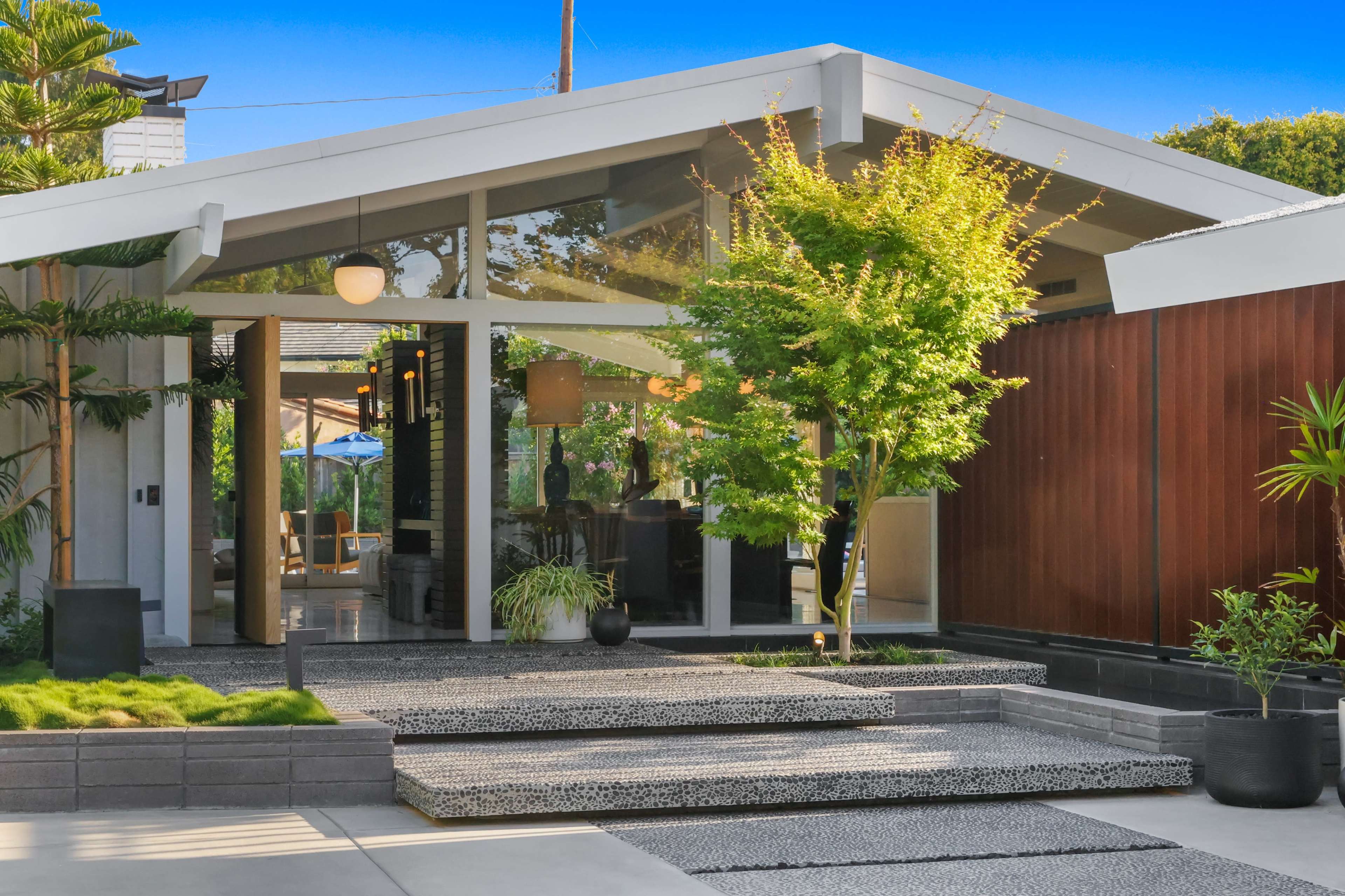 The image shows a modern house entrance with a sloped roof, surrounded by greenery and leading to a patio area.