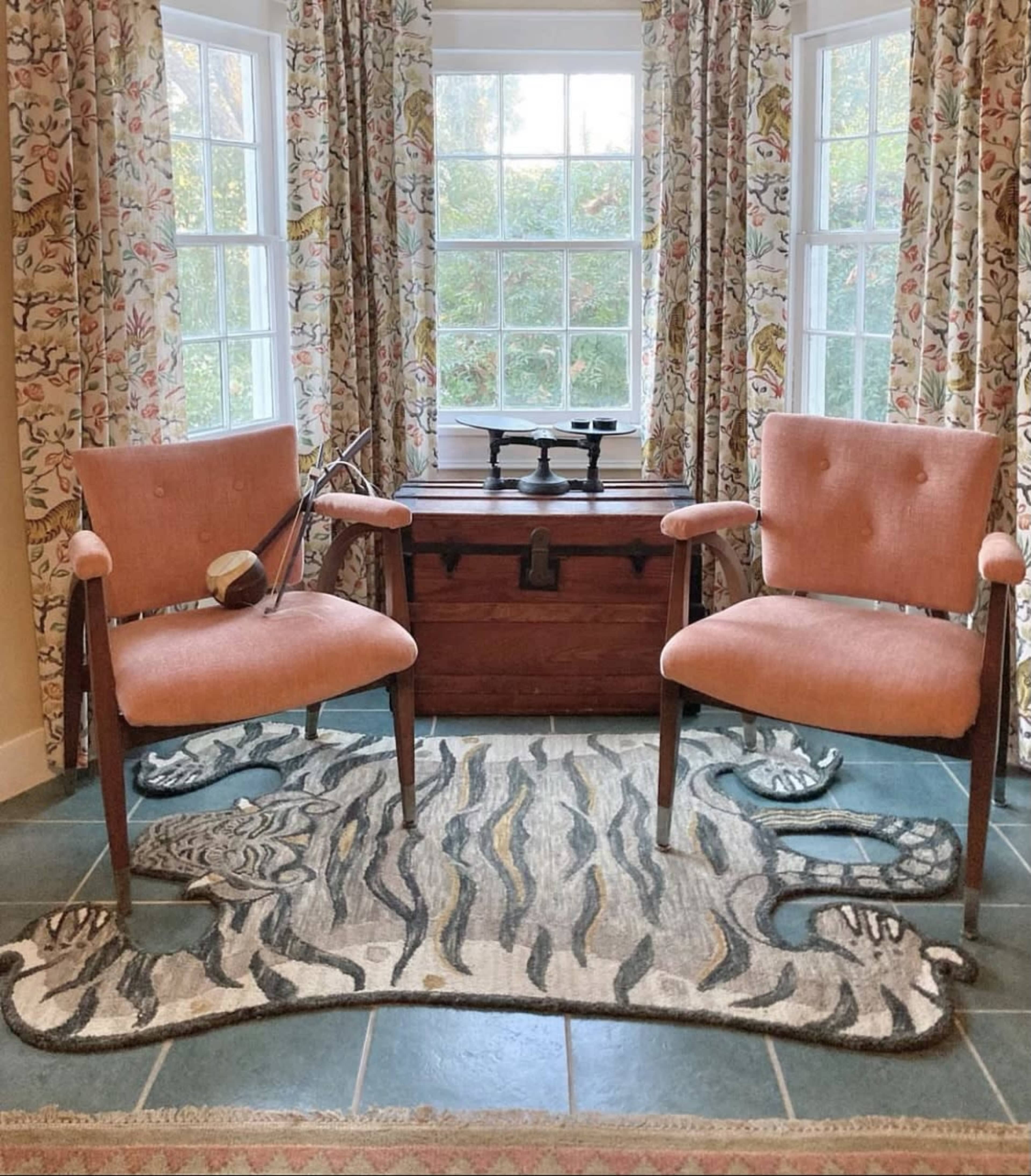 The image shows two chairs placed in front of a window, with a decorative rug underneath and a wooden trunk nearby.