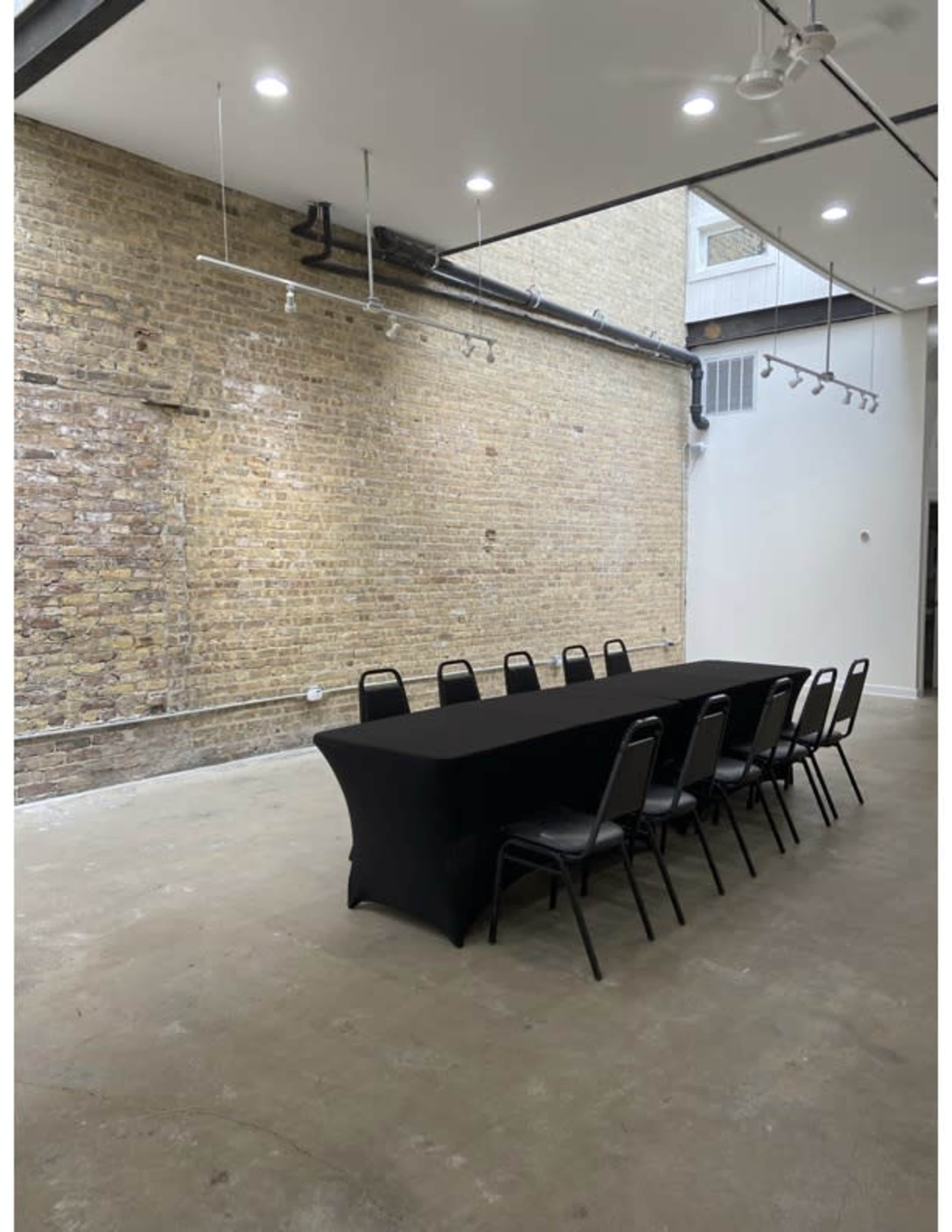 A long table with black tablecloth and metal chairs is positioned in a sparsely decorated room with exposed brick walls and concrete flooring.