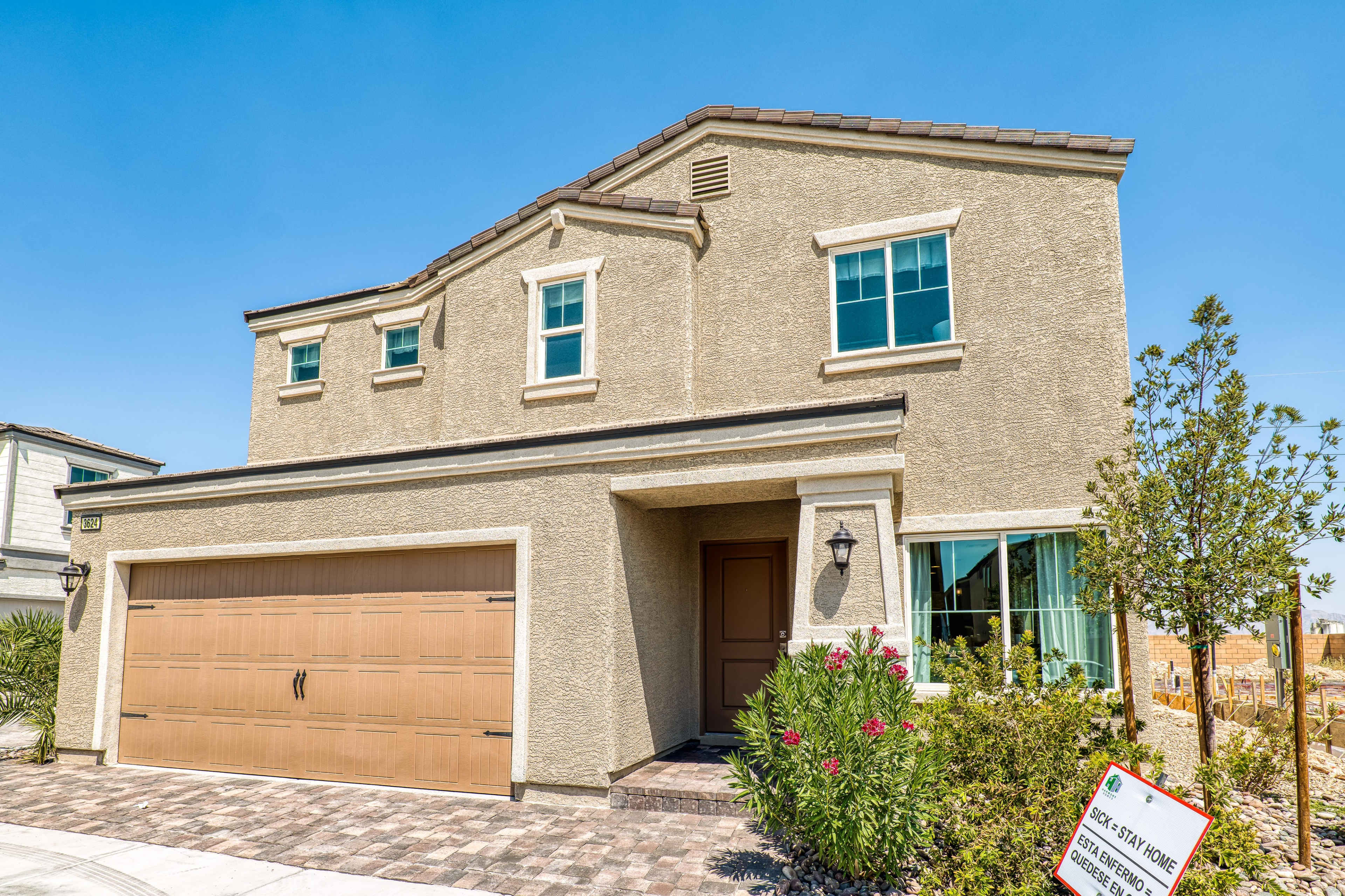 A two-story beige house with a brown garage door and large windows is situated on a paved driveway.