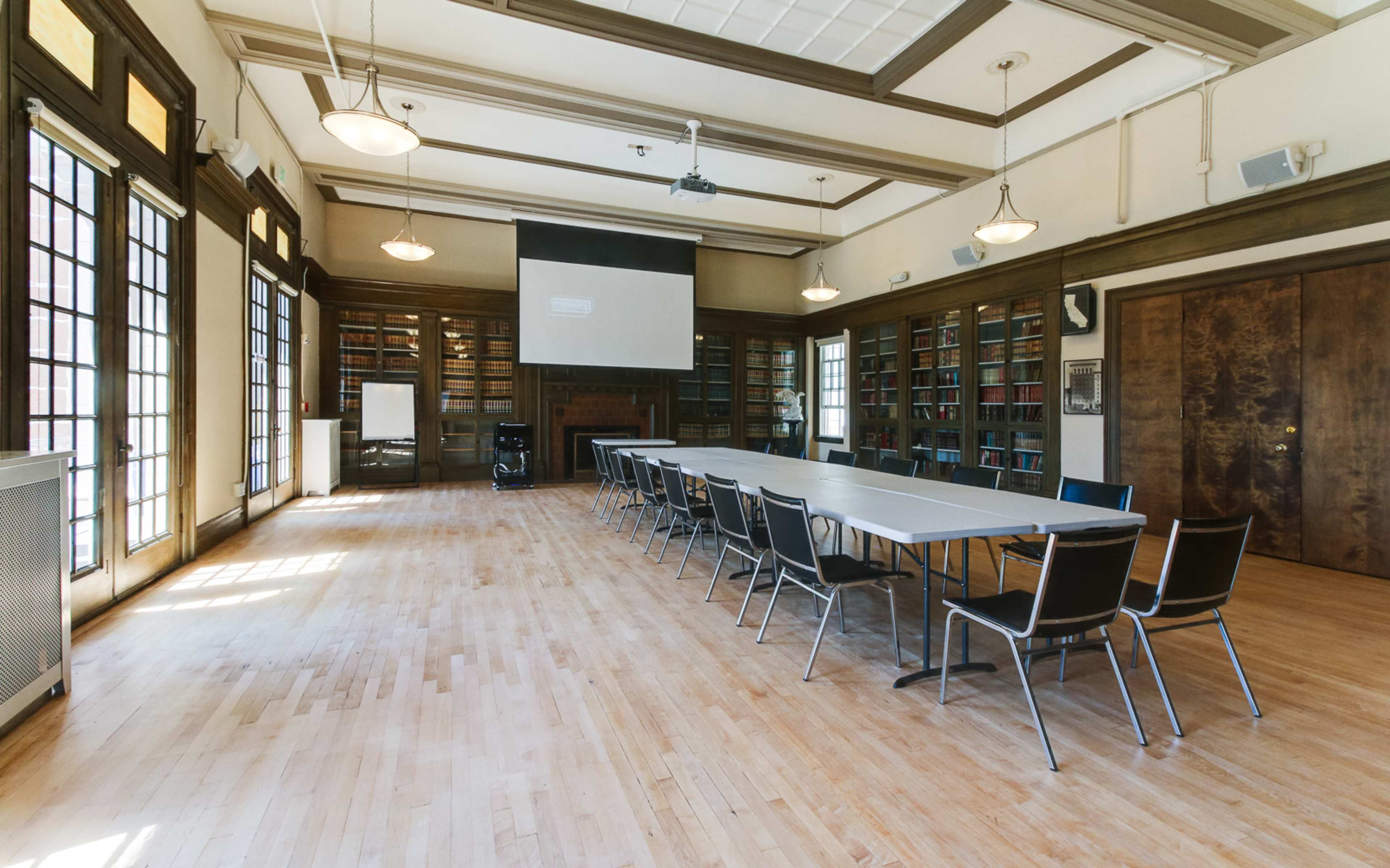 A spacious meeting room features a long table surrounded by chairs, with large windows illuminating the wooden interior and shelves filled with books.