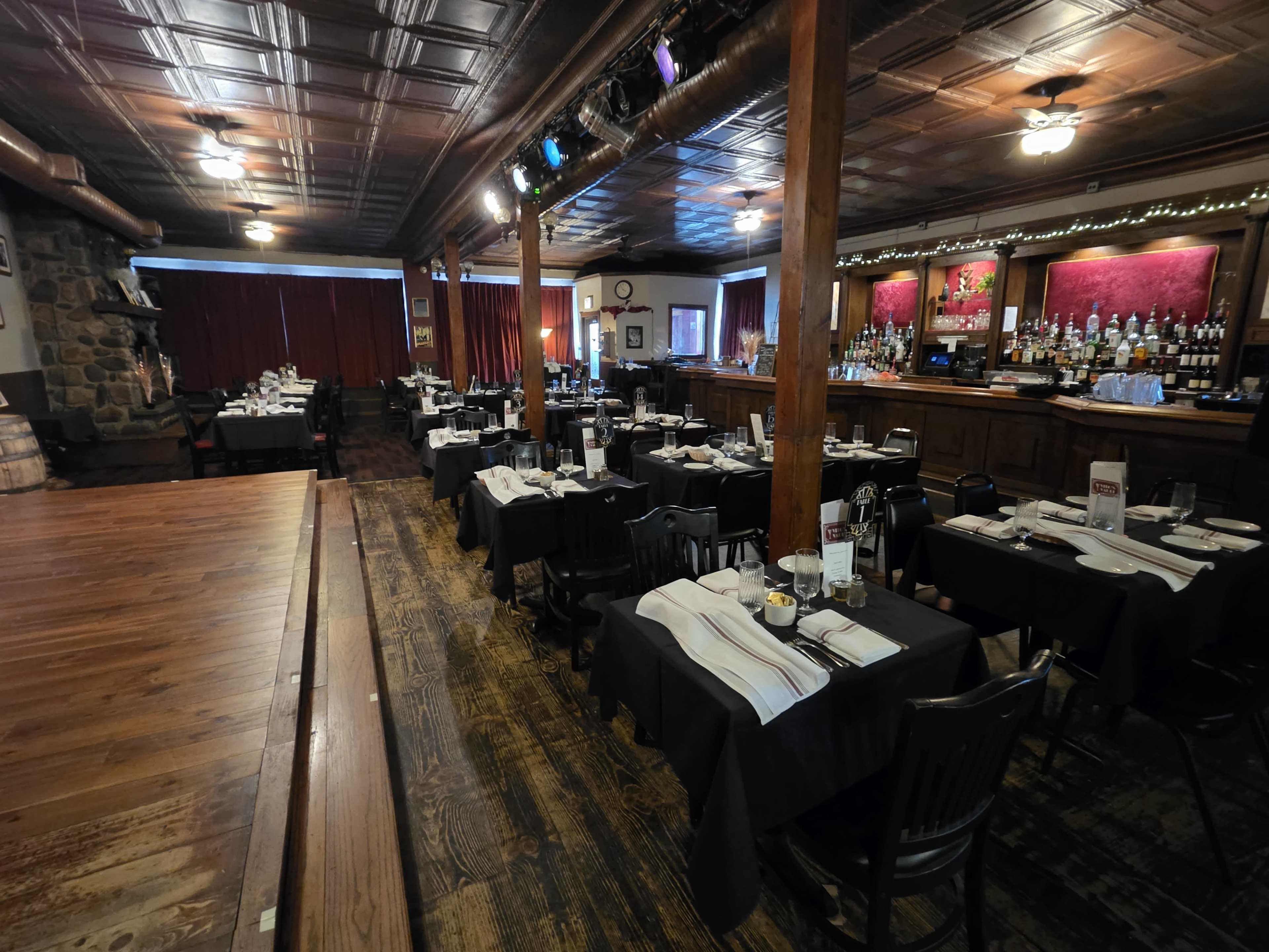 The restaurant features a spacious dining area with black tablecloths and a wooden stage, illuminated by overhead lights, and a bar area lined with bottles in the background.