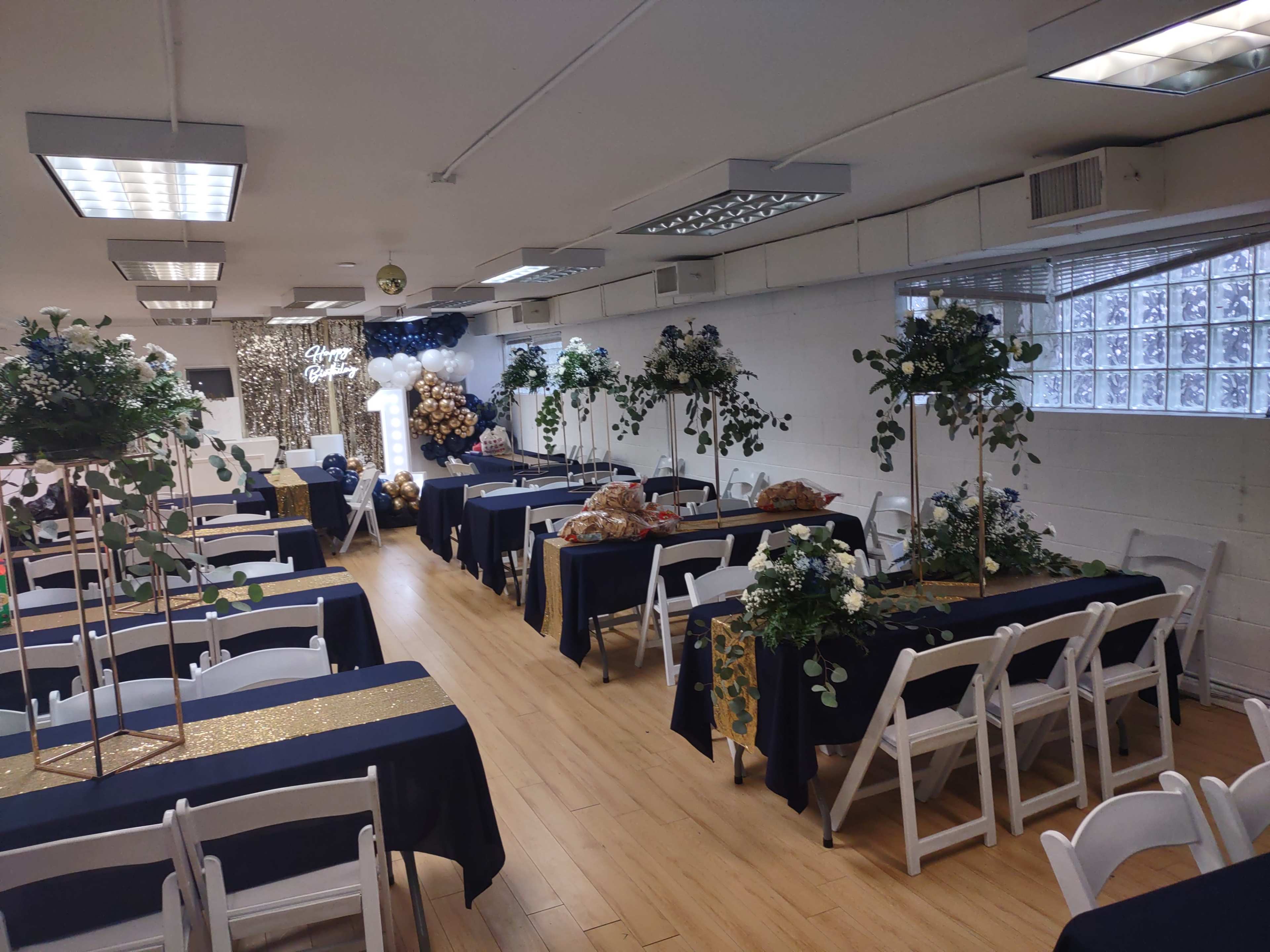 A decorated indoor event space with tables set up for a gathering, featuring blue and gold tablecloths, floral arrangements, and a backdrop with balloons.