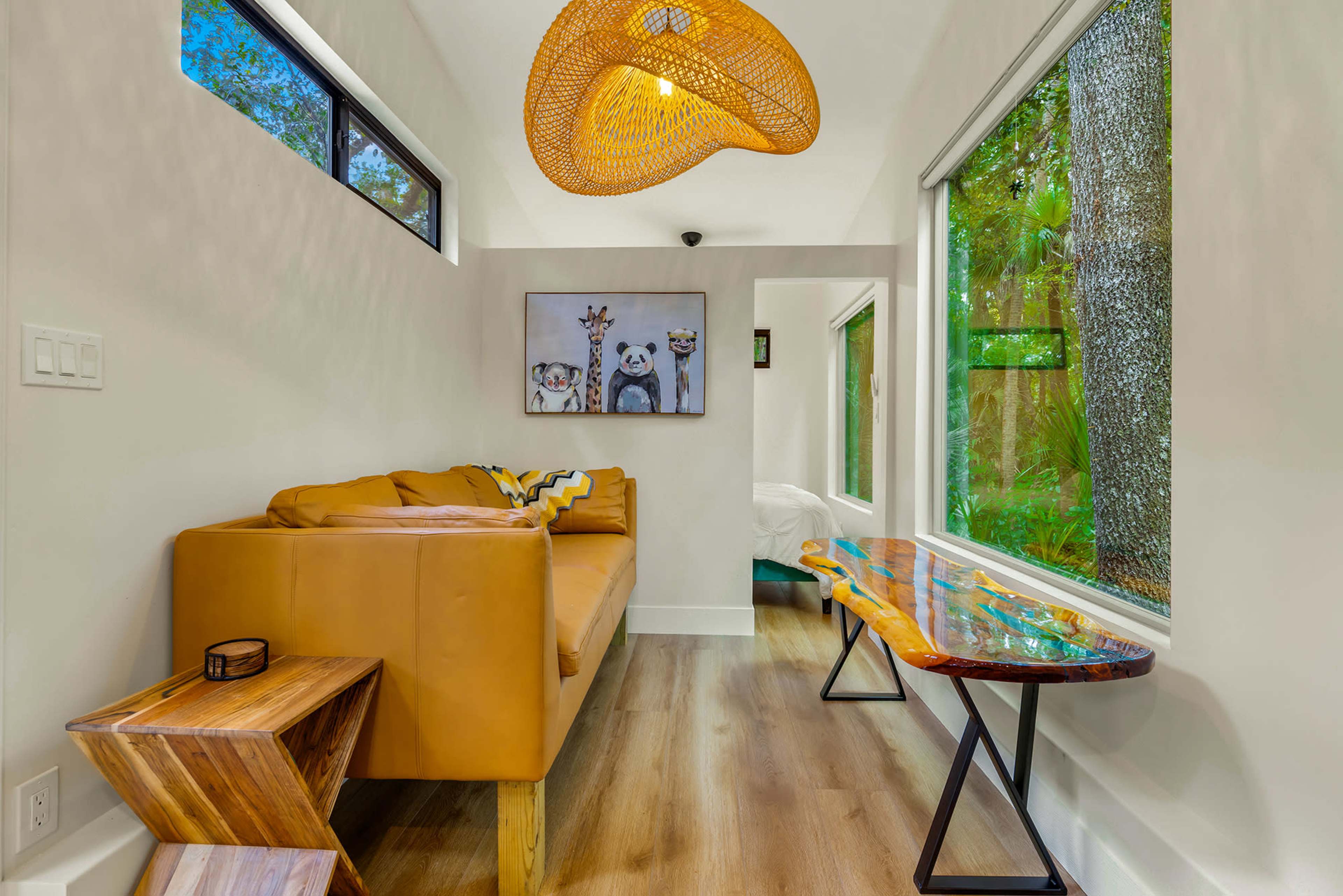 The image shows a cozy living space featuring a yellow couch, a unique wooden table, and large windows with a view of greenery outside.