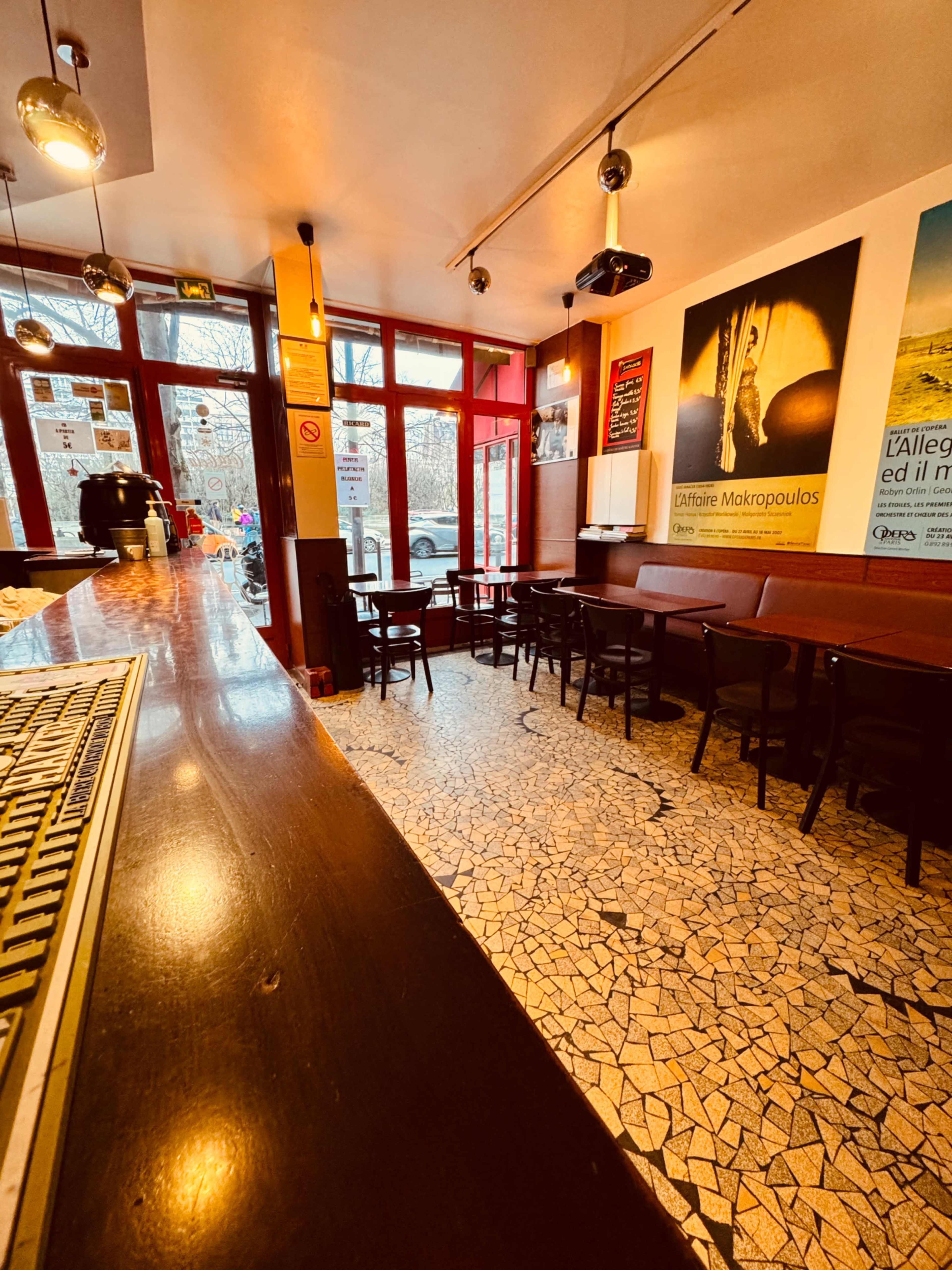 The image shows the interior of a cafe with a largely empty seating area, a long bar counter, and large windows allowing natural light to enter.