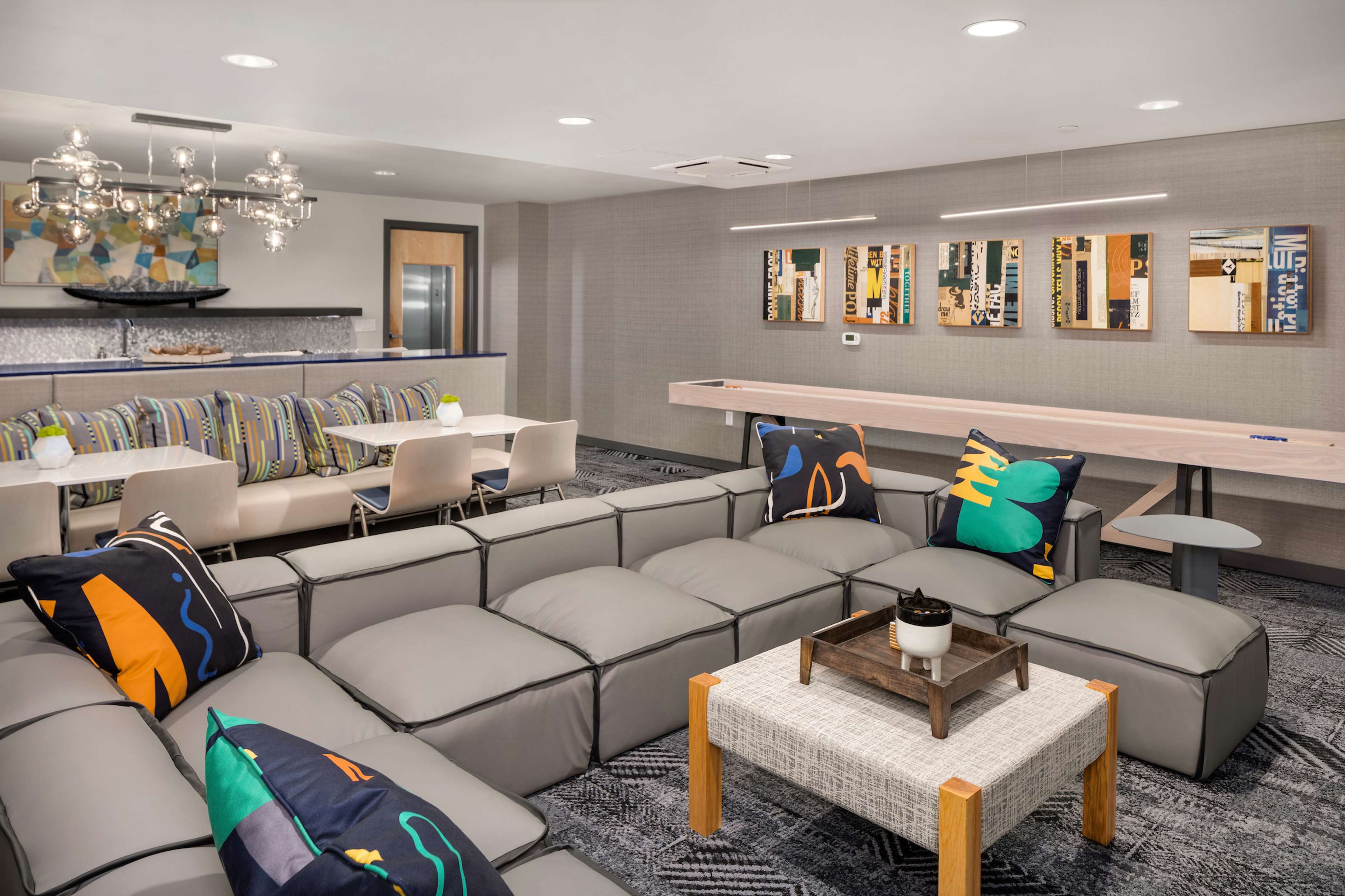 A modern communal lounge area featuring a sectional sofa with decorative pillows, a dining space with striped chairs, and a long table with wall-mounted artwork.