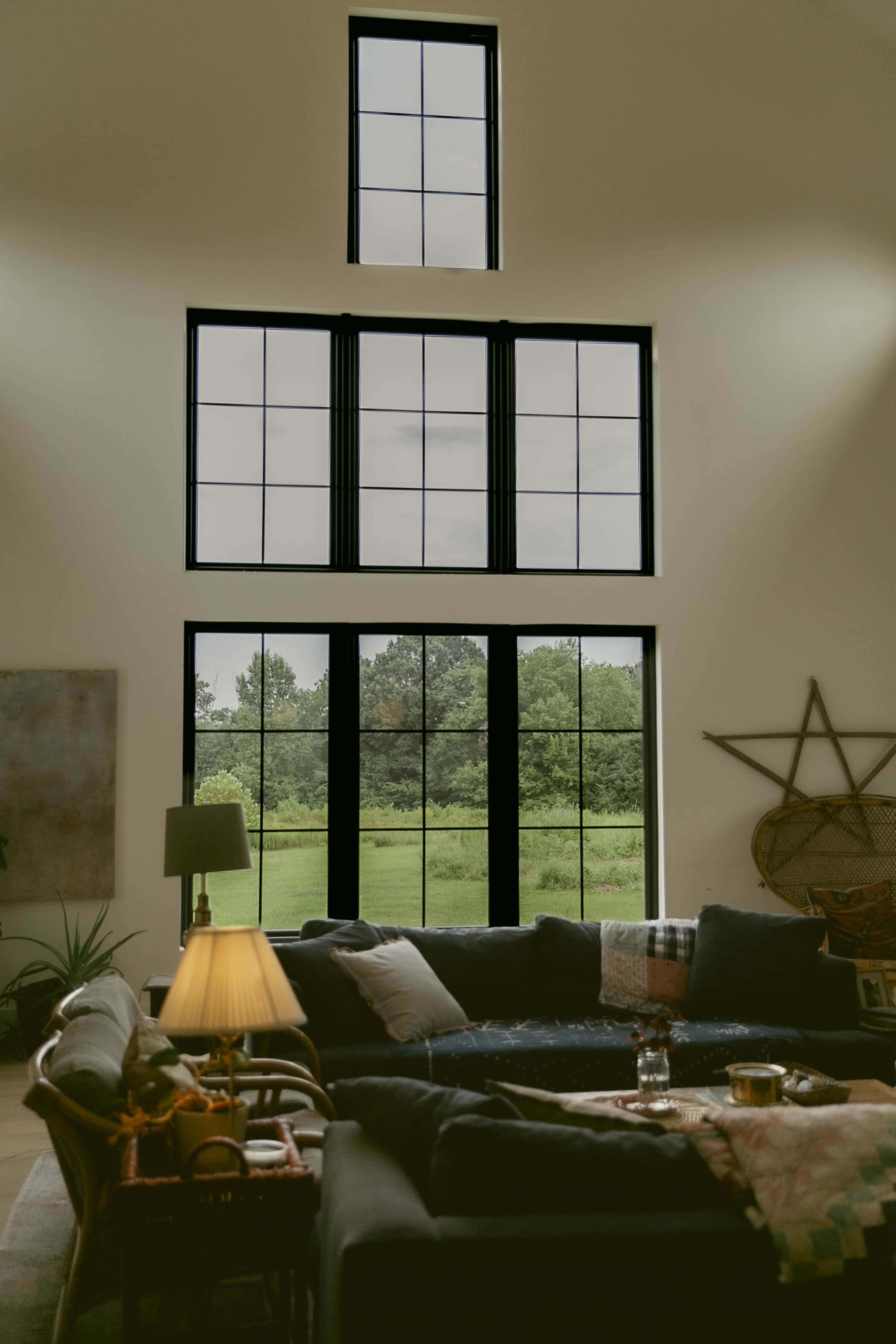 A spacious living room features a large window with multiple panes, overlooking a green landscape, and is furnished with a dark sectional sofa and a lamp.