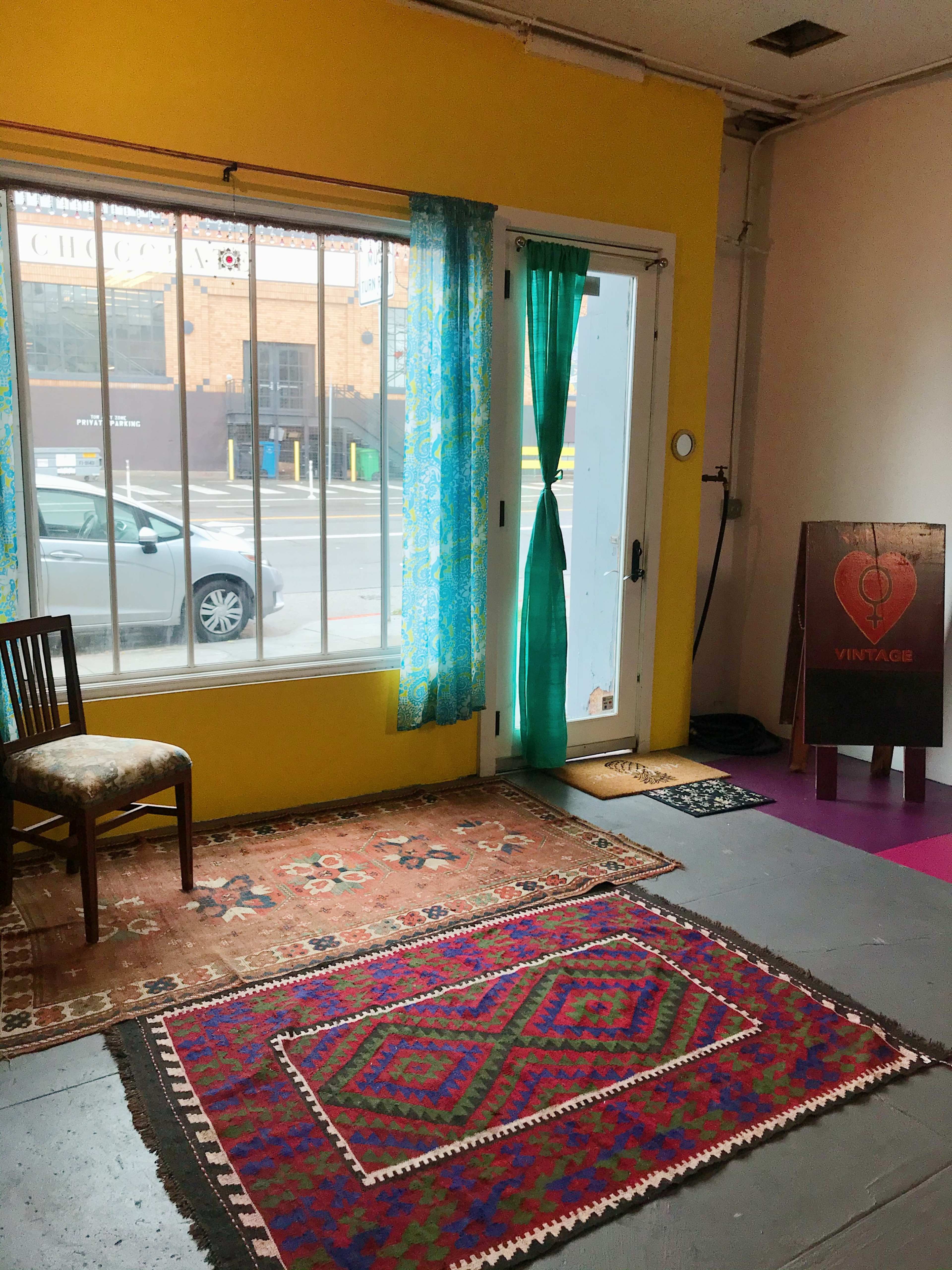 An interior space with yellow walls, a chair, colorful rugs on the floor, and a door with turquoise curtains leading to the street outside.