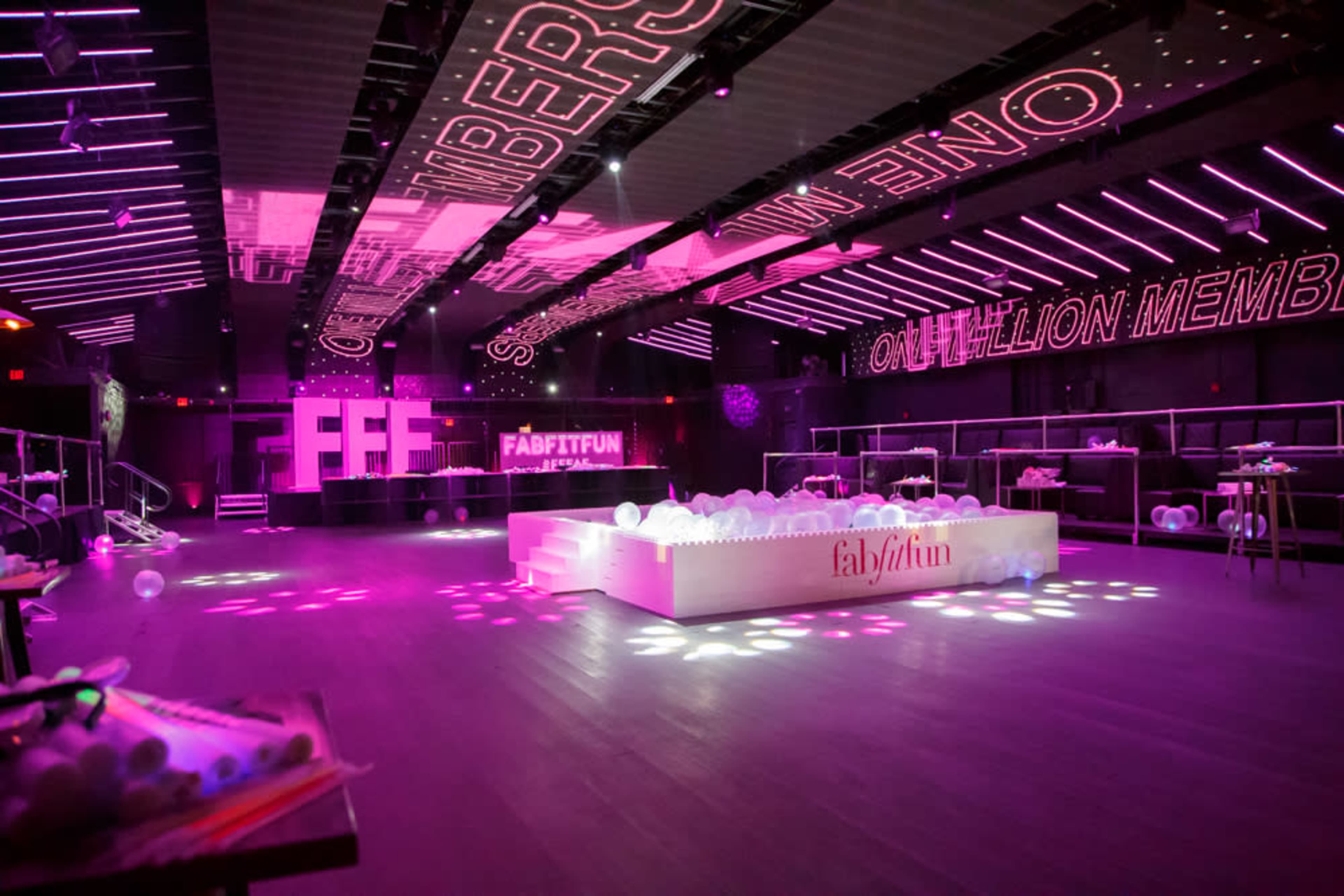 The image shows a brightly lit indoor event space with pink and purple lighting, featuring promotional displays and a central stage area.