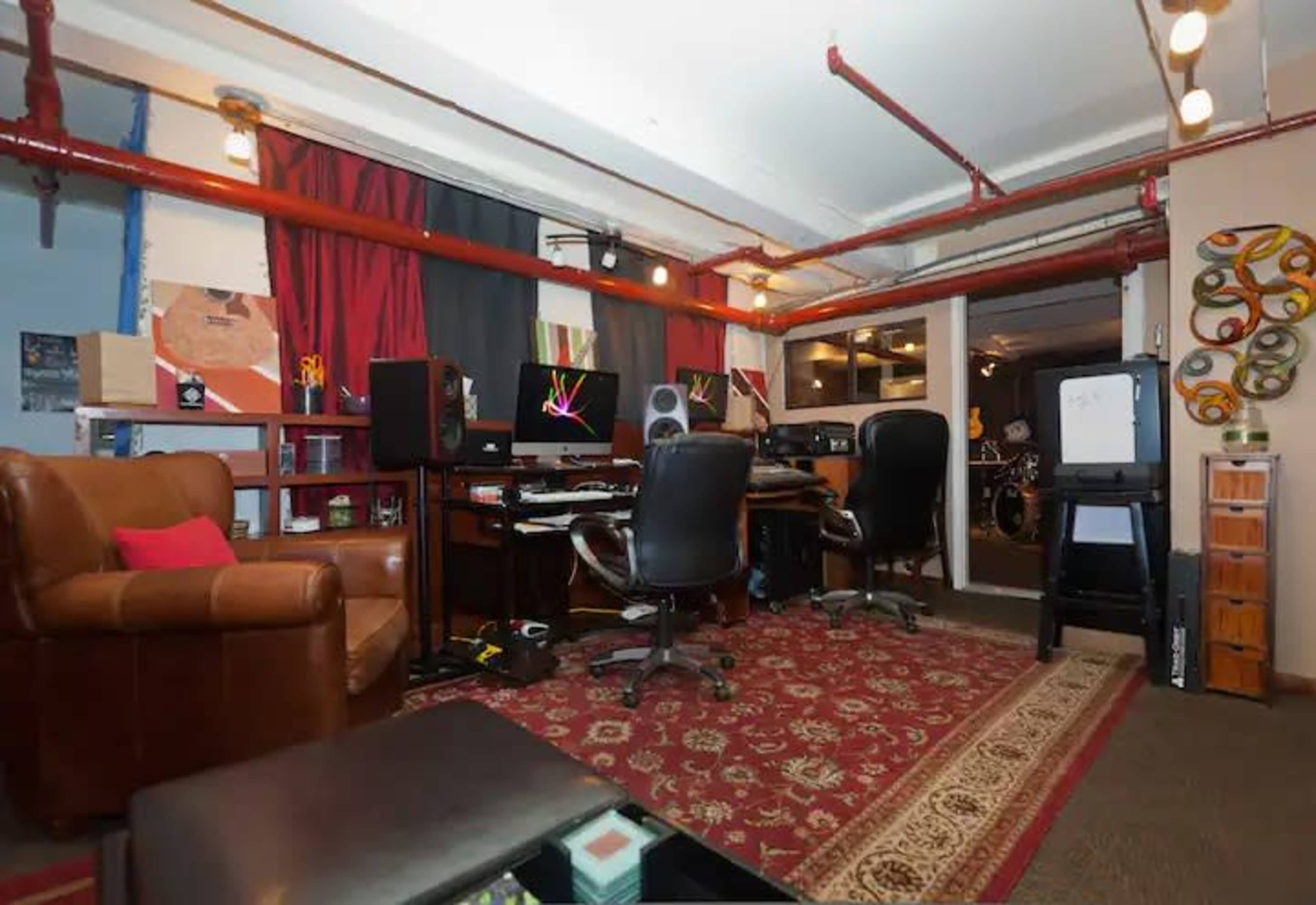 A small studio room with a desk setup featuring multiple computers, speakers, and chairs, along with a patterned rug and shelves containing various items.