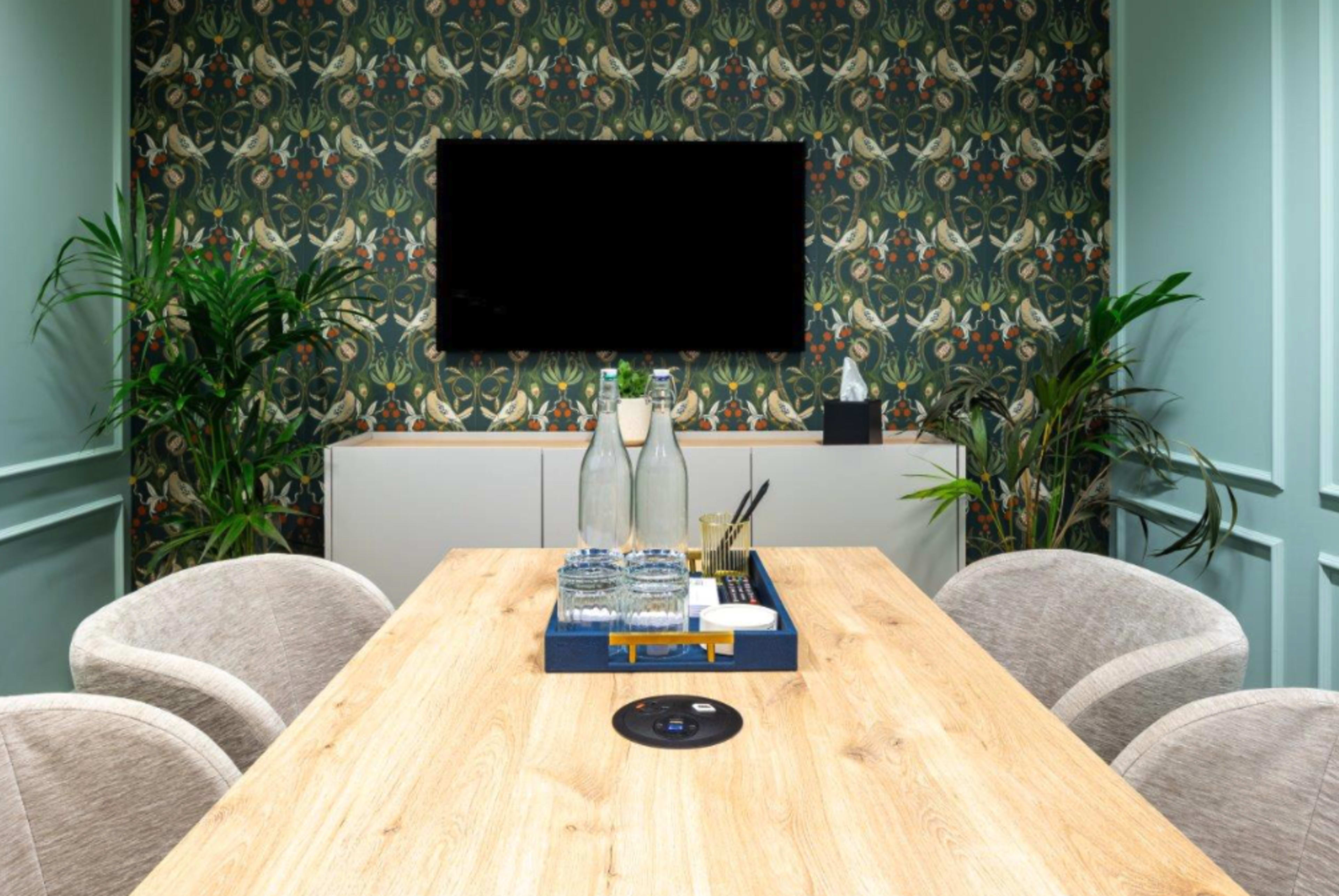 A modern meeting room features a wooden table with a decorative tray, surrounded by upholstered chairs, and is adorned with floral-patterned wallpaper and a large blank television screen.
