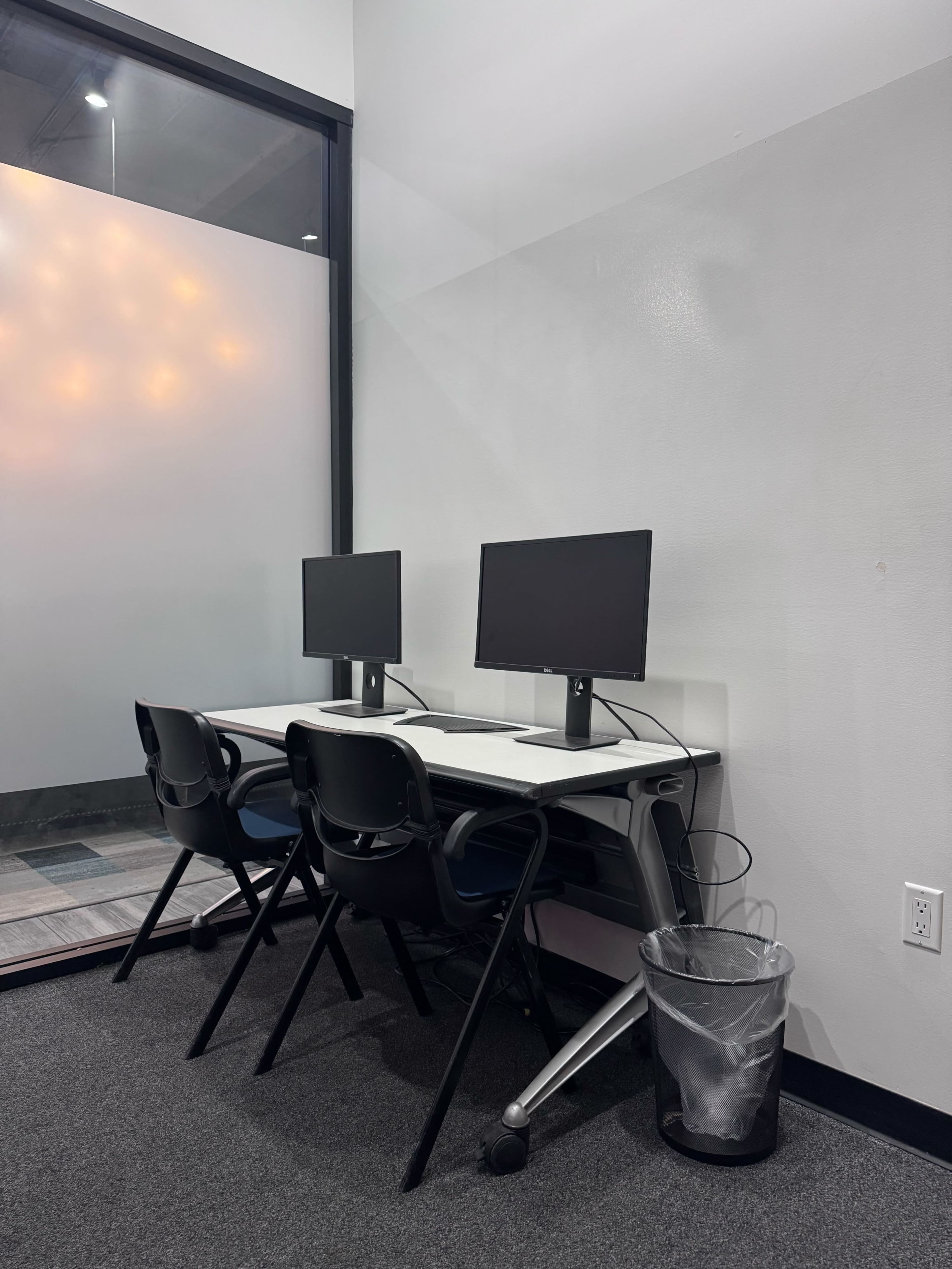 The image shows a small workspace with two computer monitors on a white desk and three black chairs, next to a trash bin in a modern office setting.