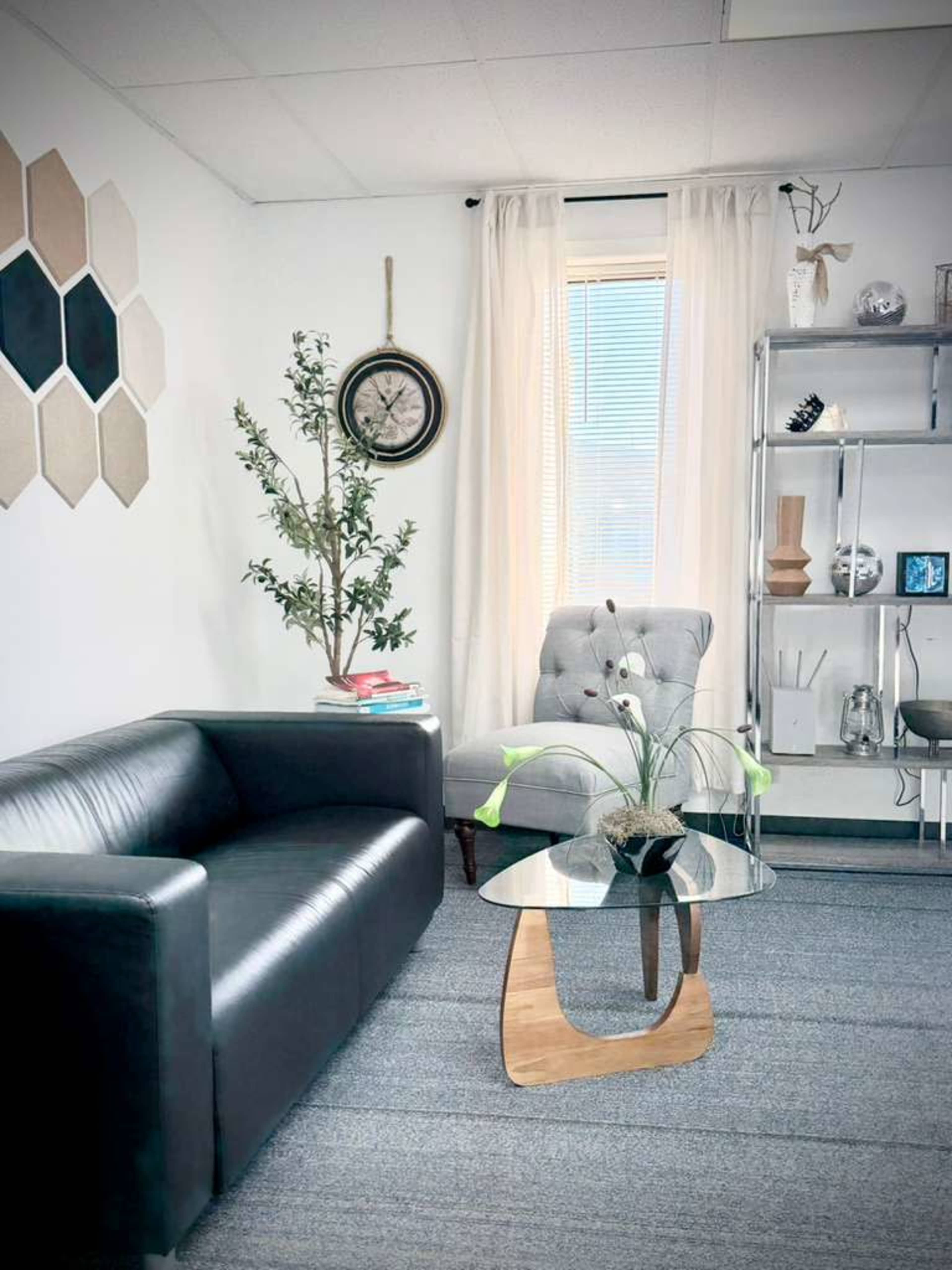 The image shows a modern office space featuring a black leather sofa, a glass coffee table, a gray tufted armchair, decorative plants, and a wall display of hexagonal art pieces.