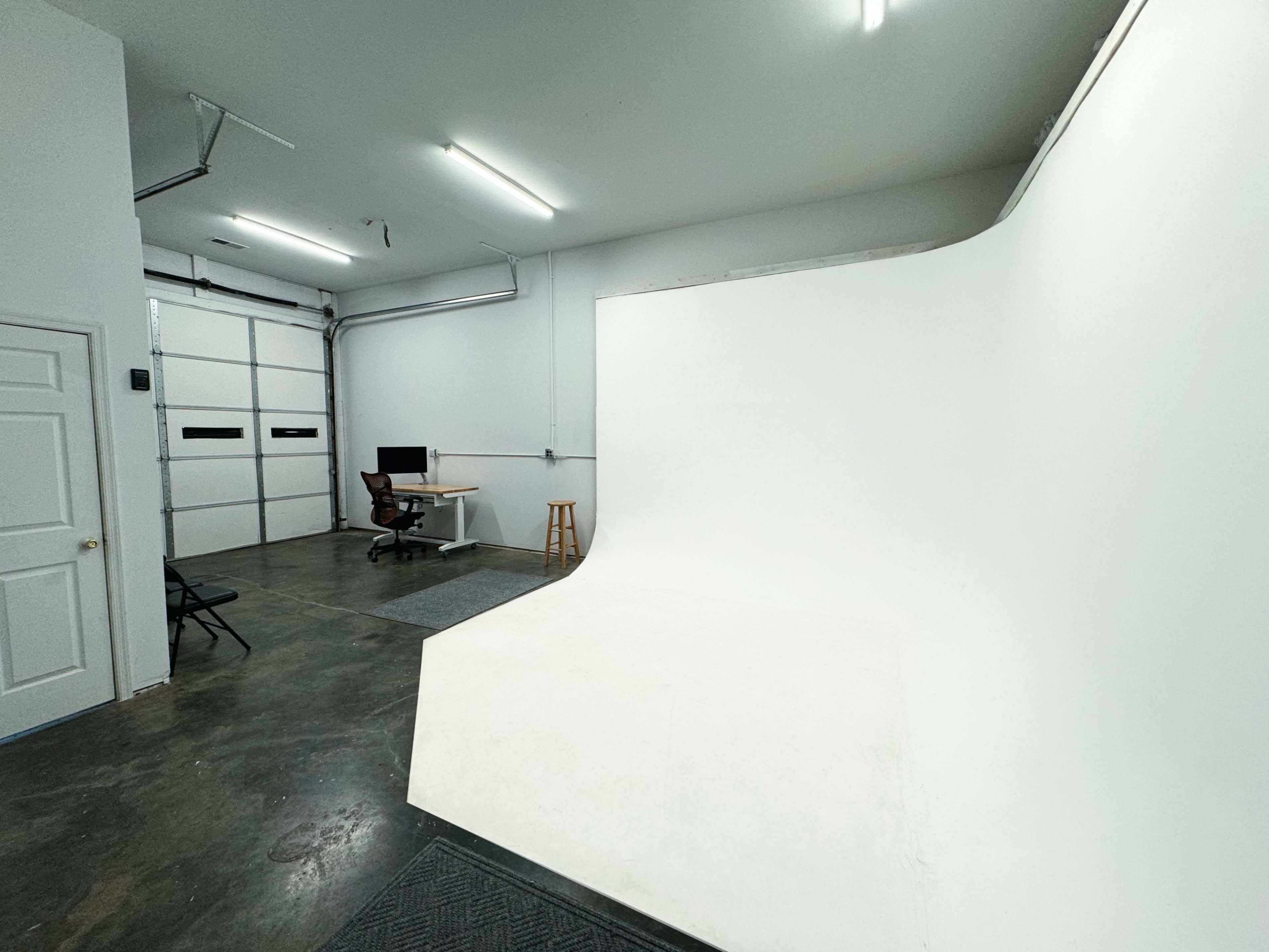 An empty garage space with a seamless white backdrop on one side and a desk with a computer in the corner.