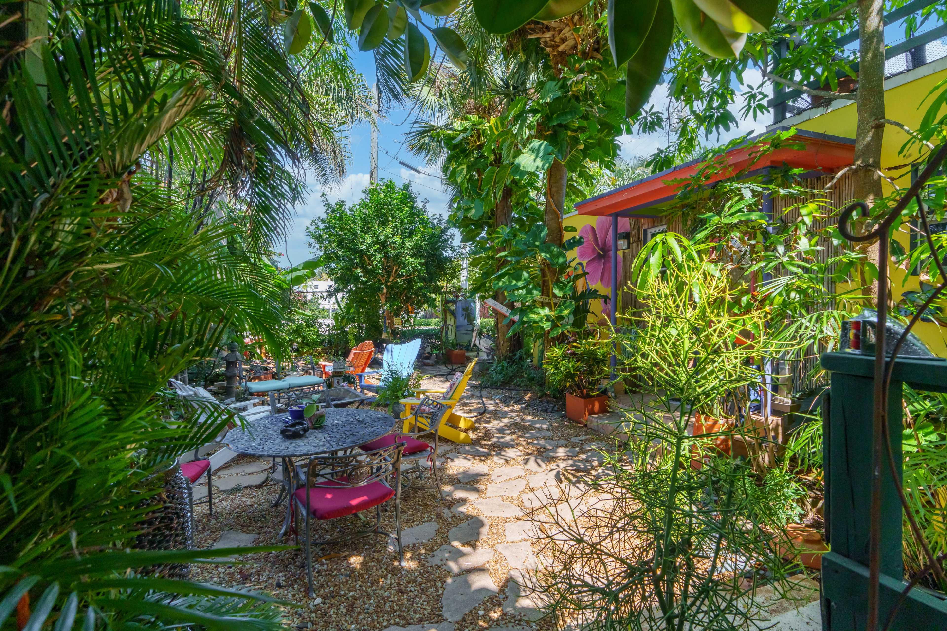 A lush garden features a variety of seating options and plants, with a stone path leading to a colorful building.