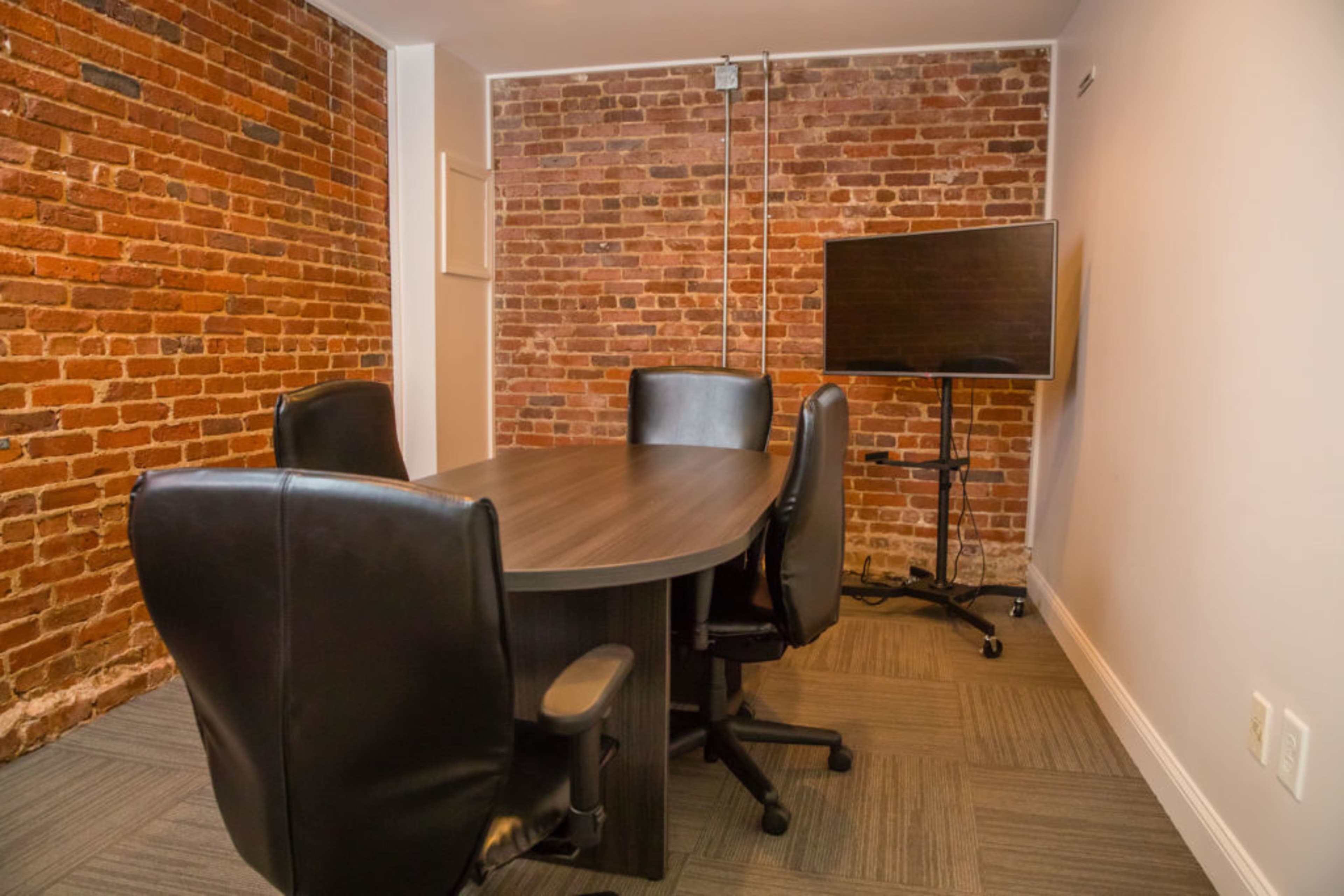 A conference room featuring a wooden table surrounded by four black chairs, with a brick wall and a mounted television.