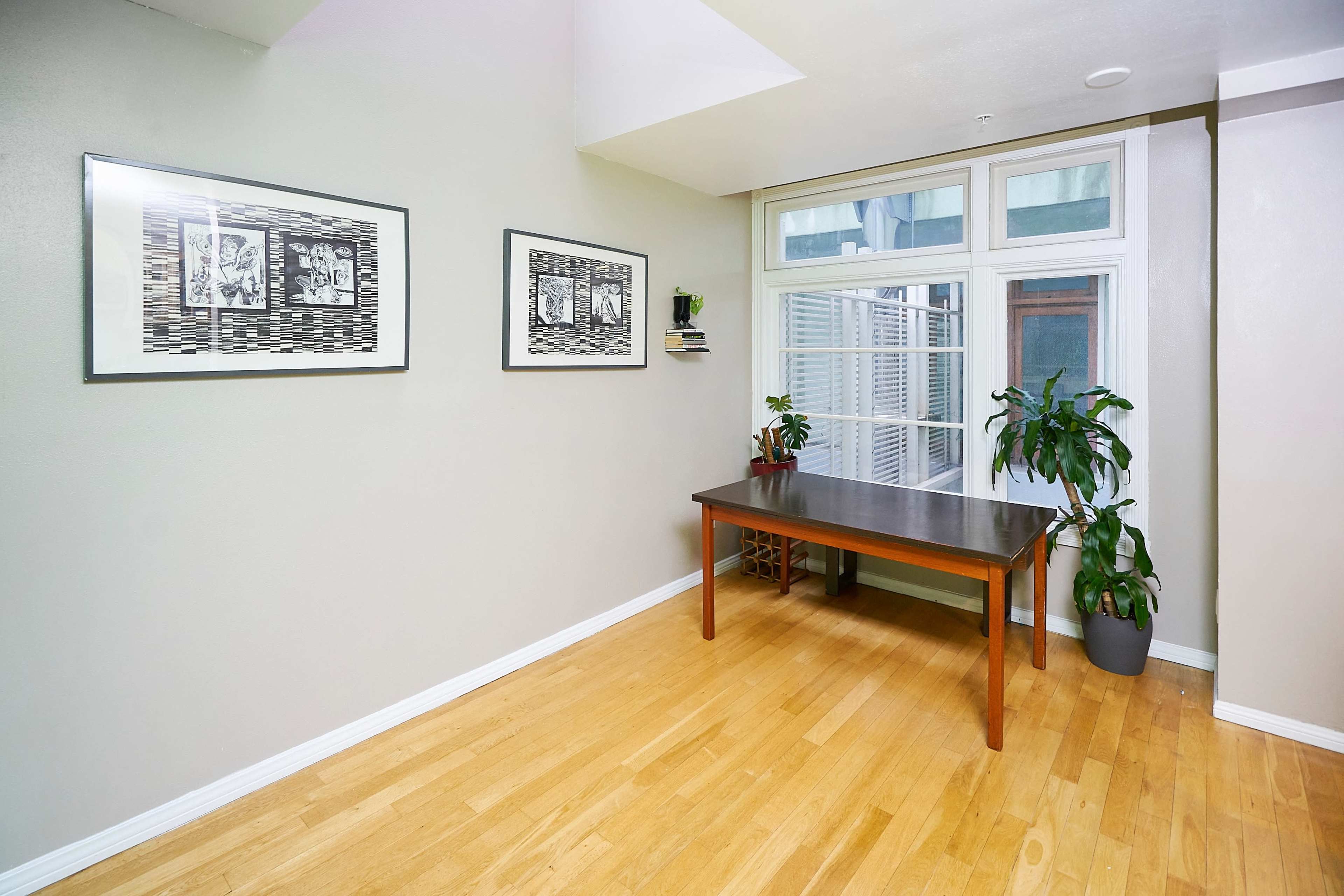 A room with hardwood flooring, a wooden table, two framed artworks on the wall, and a plant in a pot near a window.