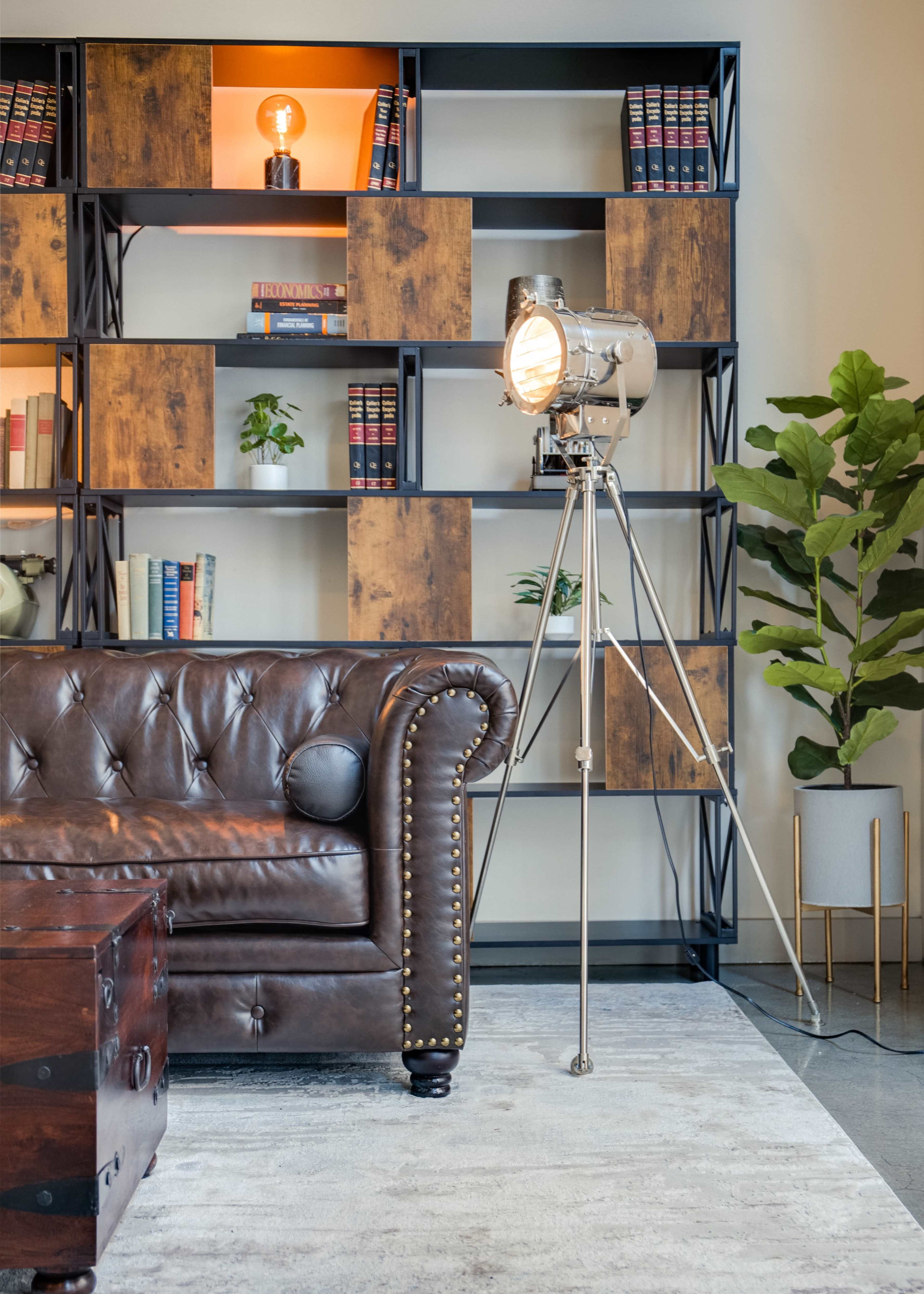 A vintage-style tripod lamp stands beside a brown leather chesterfield sofa, with a wooden bookshelf filled with books in the background.