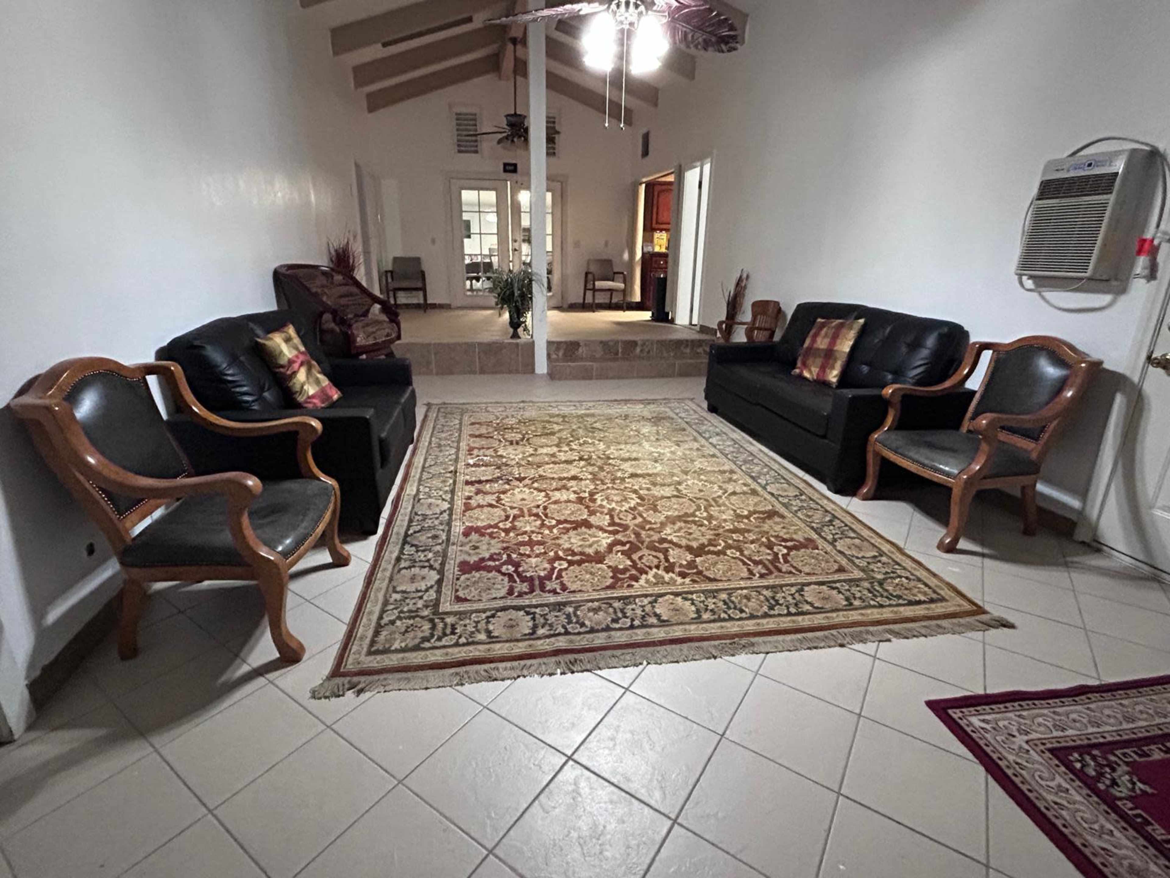 The image shows a spacious living room featuring two black sofas and two wooden chairs arranged around a large area rug, with a view of a raised platform in the background.