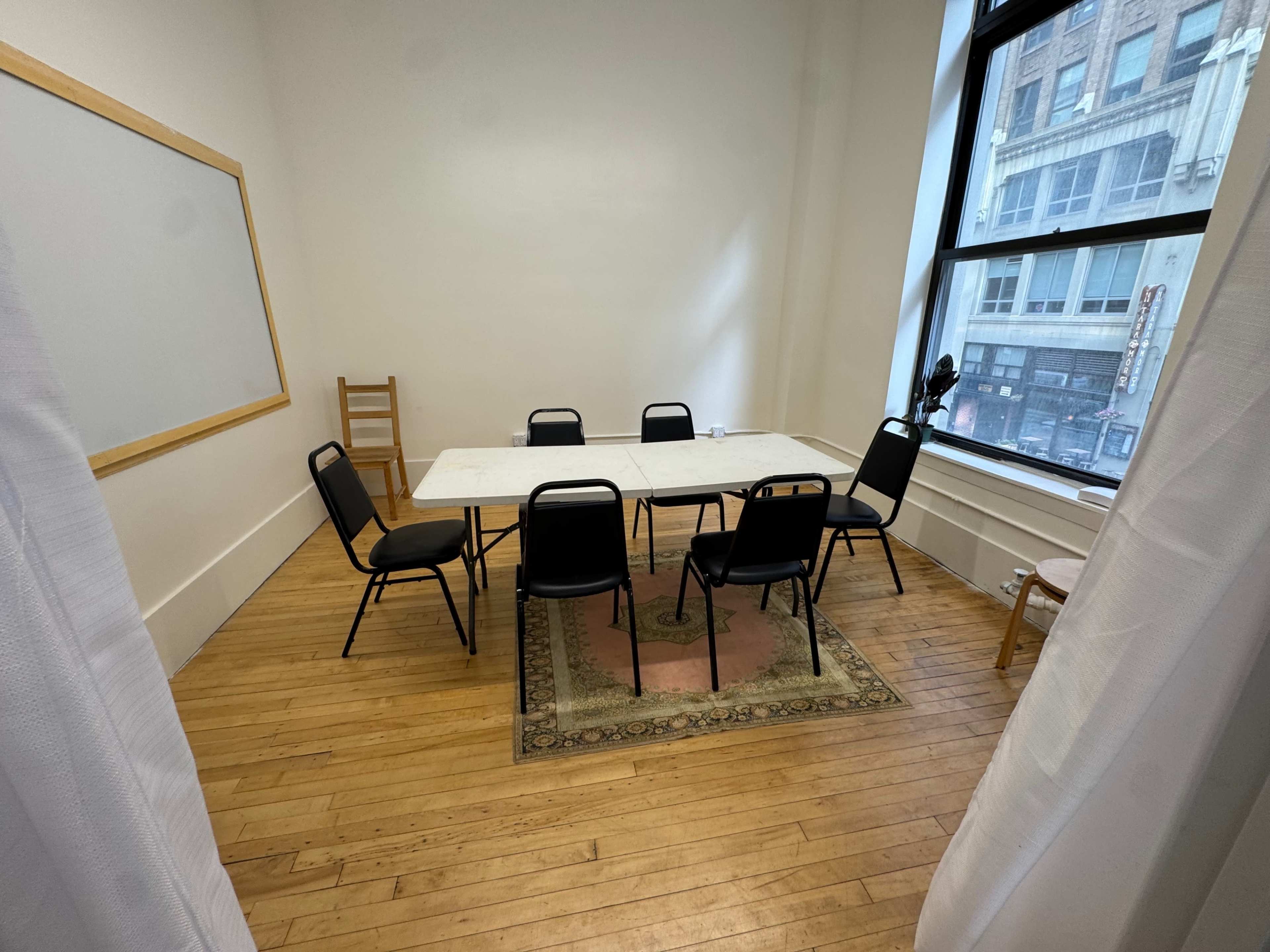 A small conference room features a rectangular table surrounded by six black chairs, with large windows allowing natural light to enter.