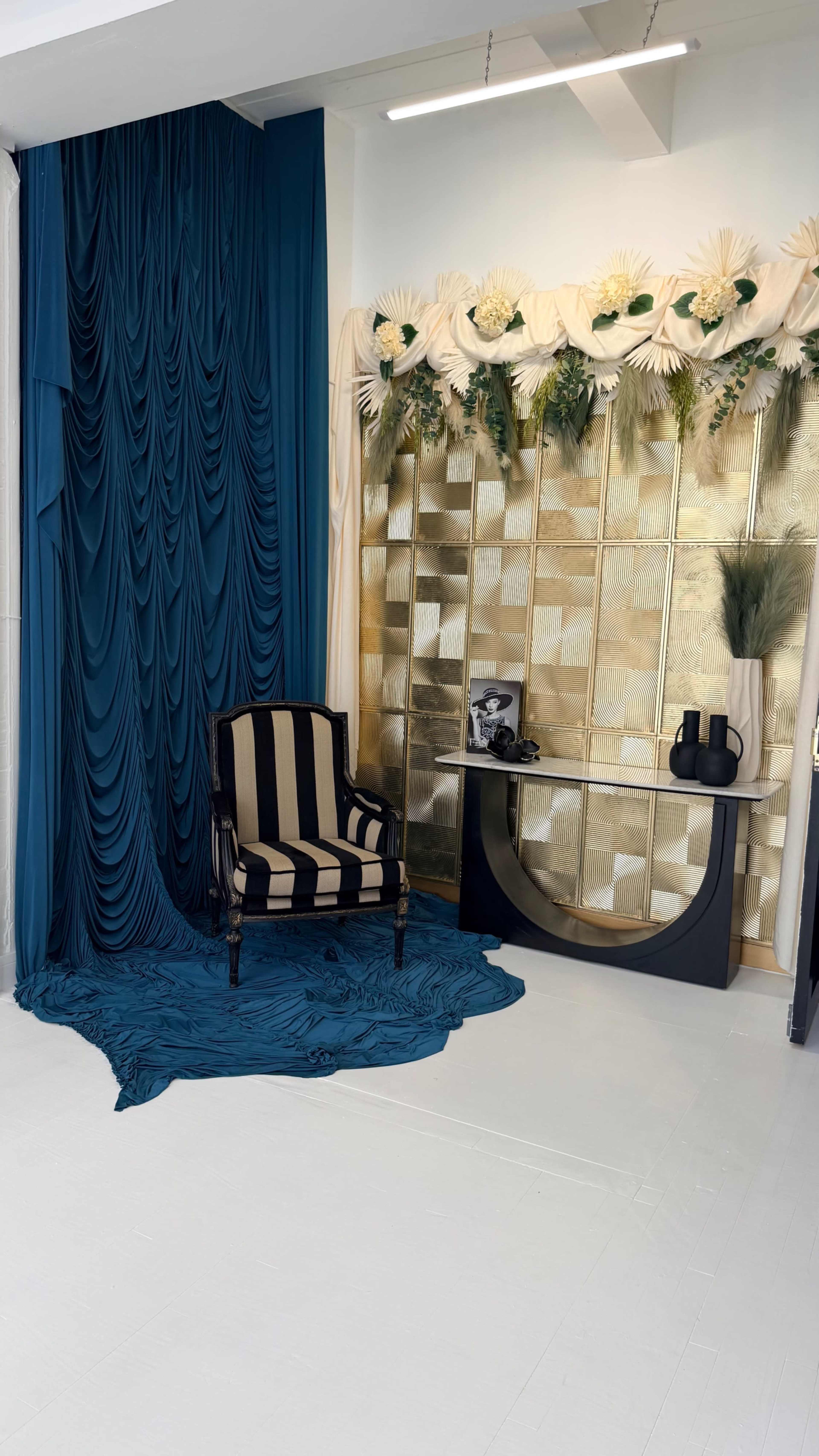 The image shows a stylish interior scene featuring a striped chair, a console table, and a backdrop of draped teal fabric and golden decorative panels.