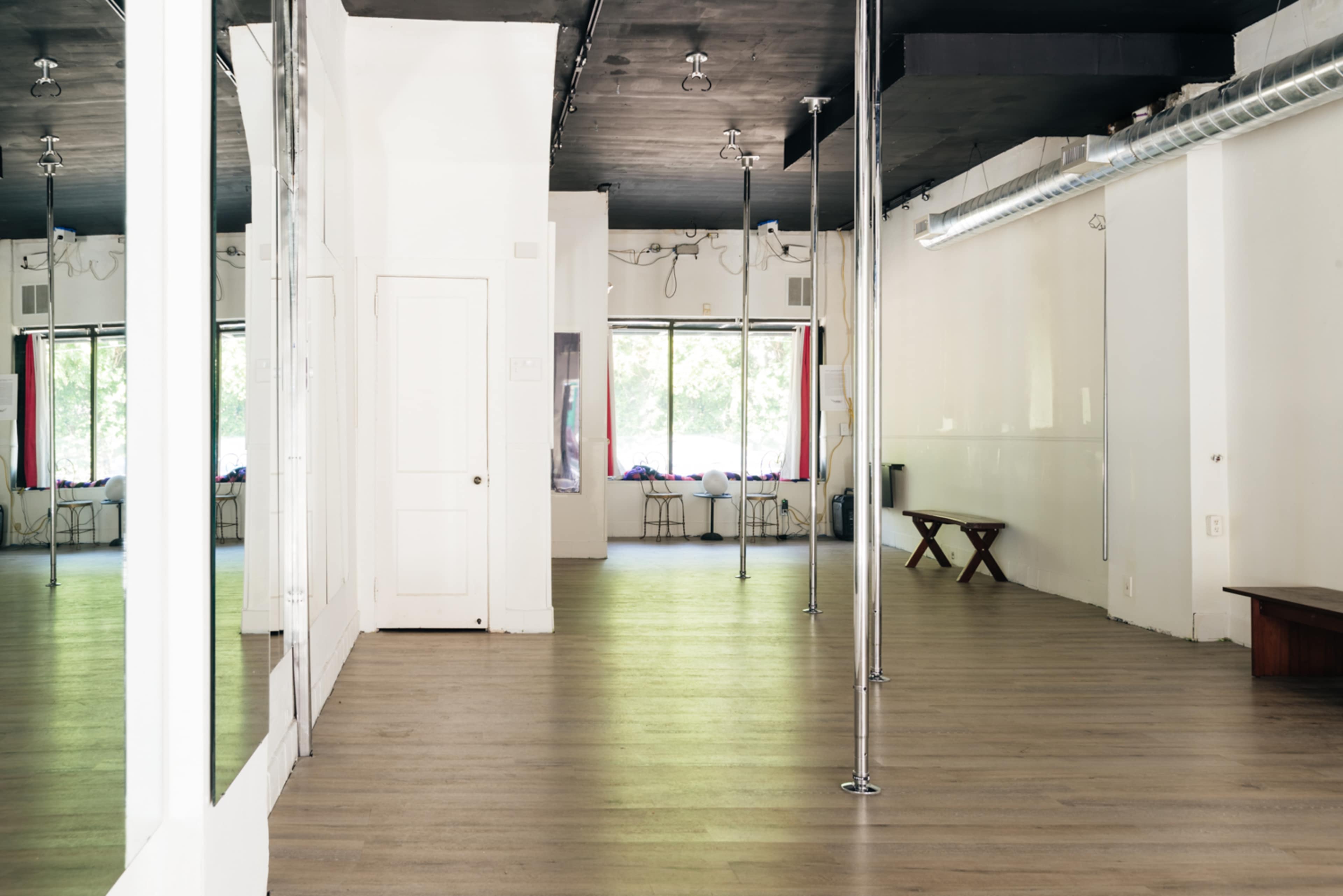 A spacious dance studio with polished wooden floors, mirrors on one wall, and poles set up for dancing.