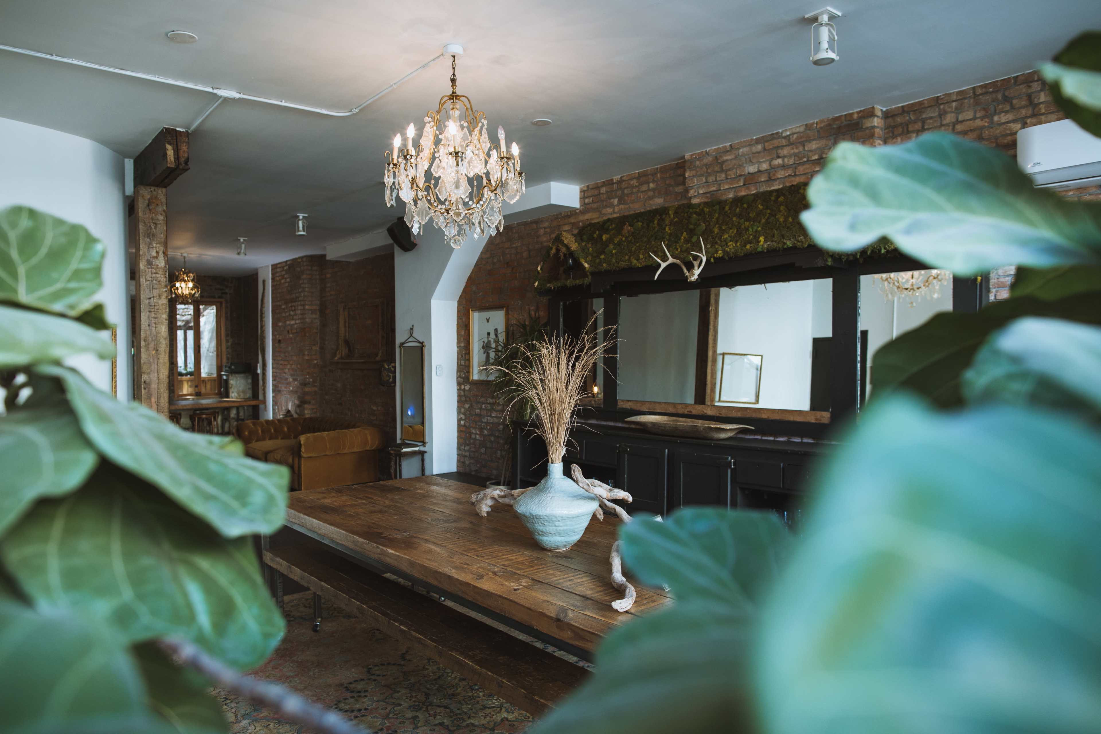 A spacious room features a large wooden table with a vase, a chandelier, and decorative mirrors against a brick wall.