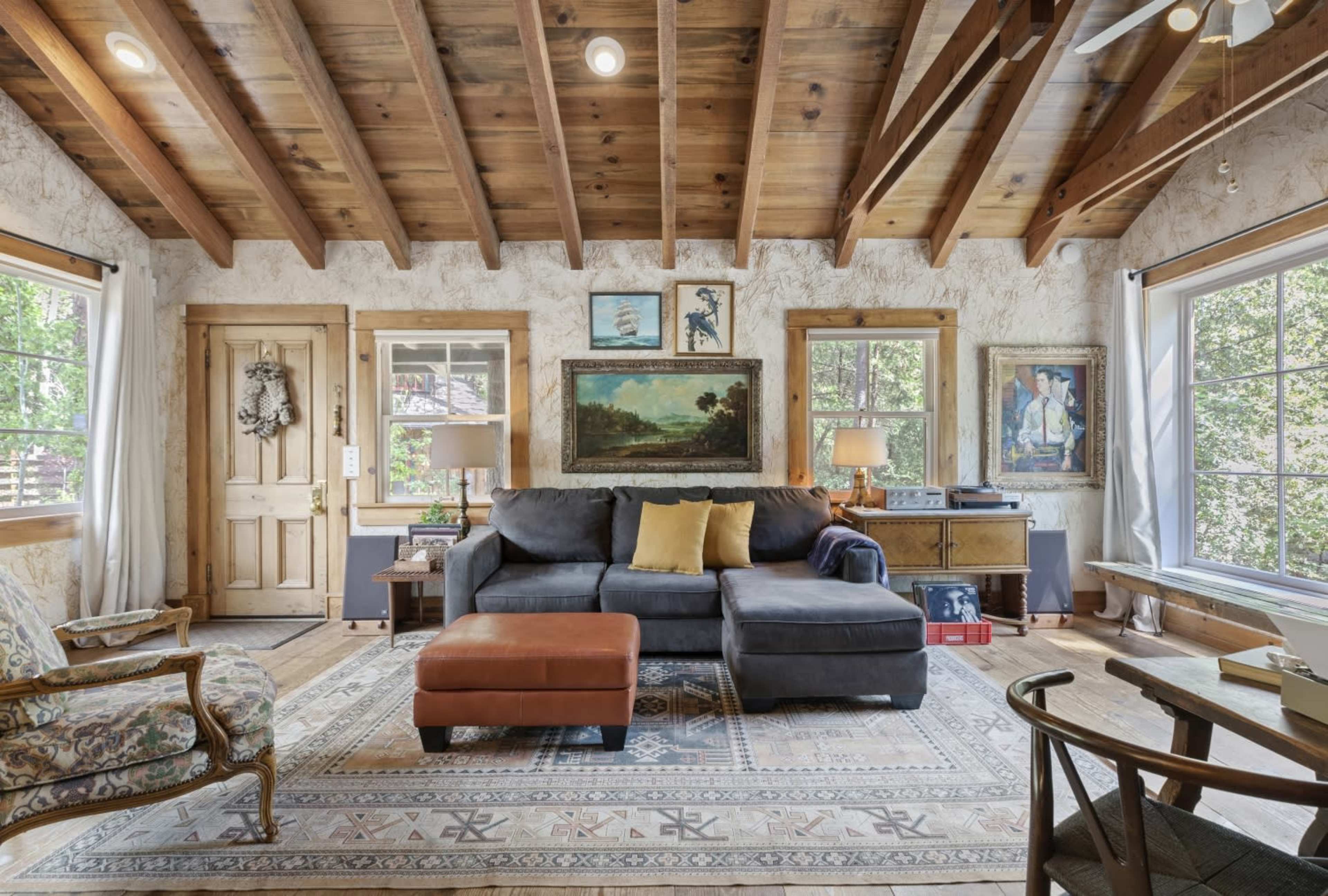 The living room features a gray sofa, a brown ottoman, wooden beams on the ceiling, and several framed paintings on the walls, with large windows allowing natural light to enter.
