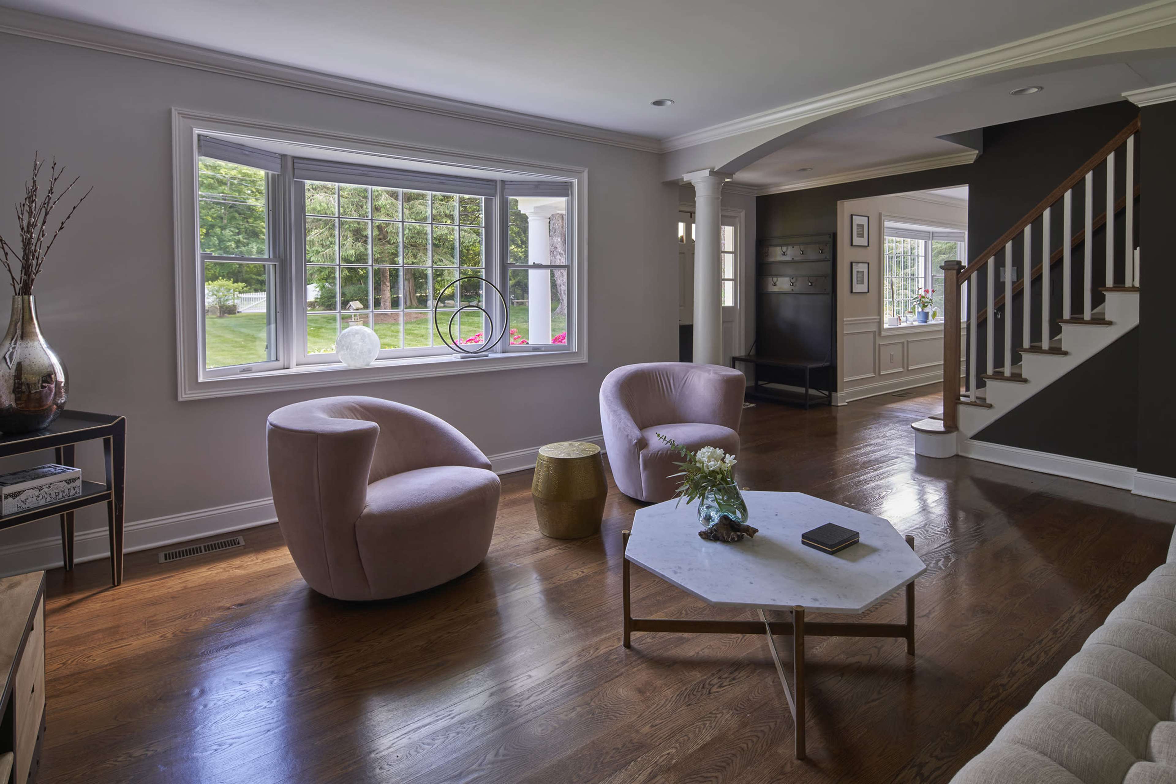 A light-filled living room with two pink chairs, a marble coffee table, and a staircase leading to another room.