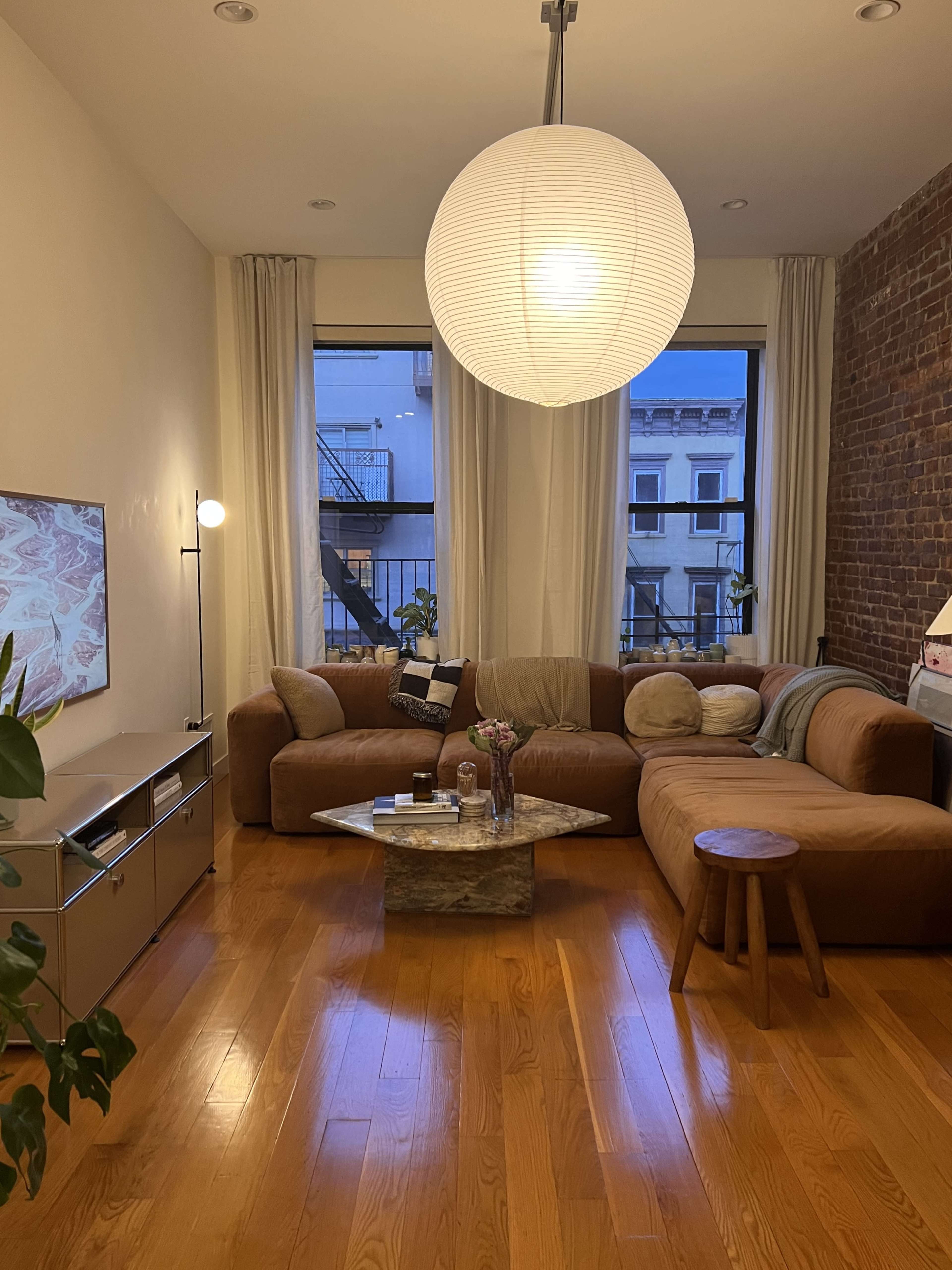 A living room features a large brown sectional sofa, a wooden coffee table, and a pendant light, with large windows allowing natural light to enter.