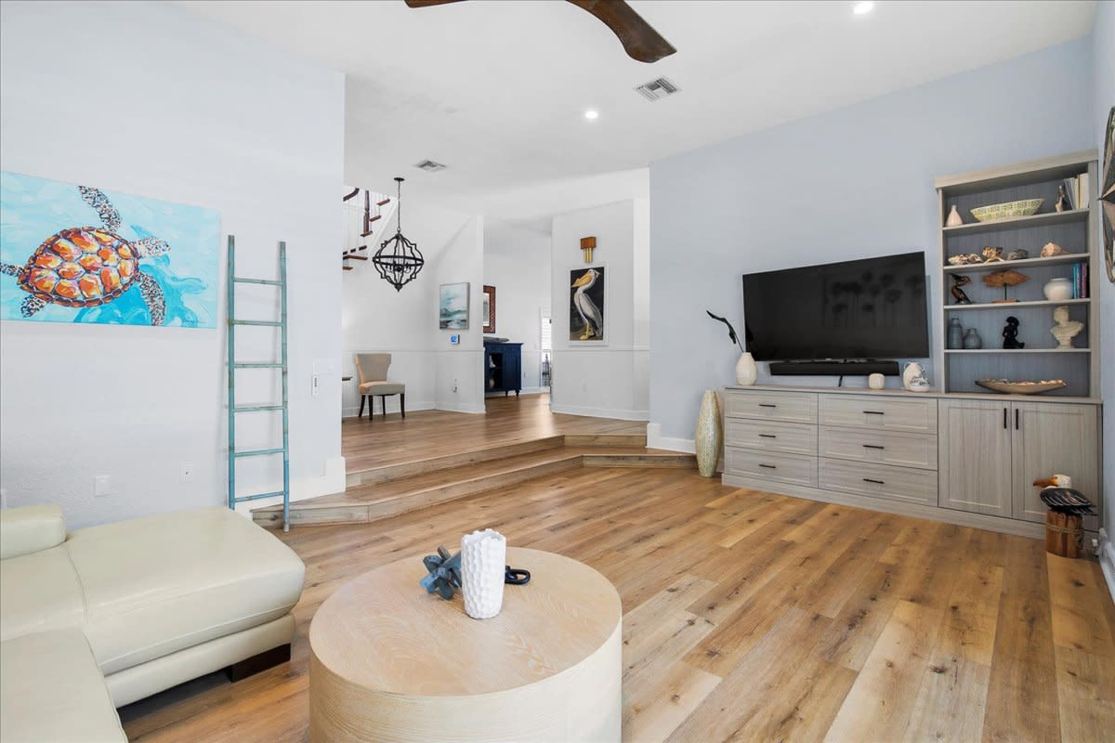 The image features a spacious living room with a light-colored sectional sofa, wooden flooring, a mounted television, and decorative shelves against a soft blue wall.