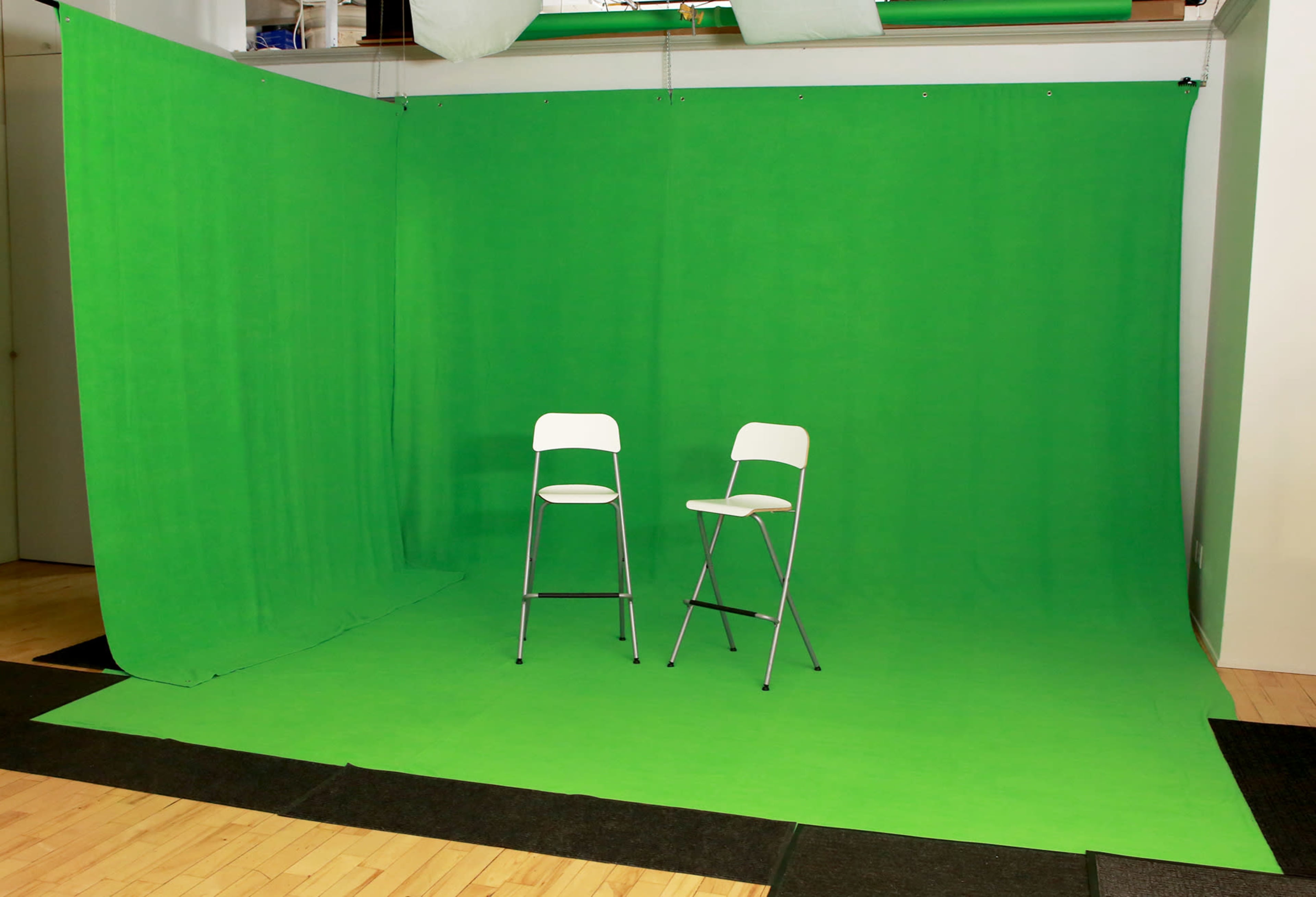 The image shows a photography studio with a green backdrop and two metal stools positioned in front of it.