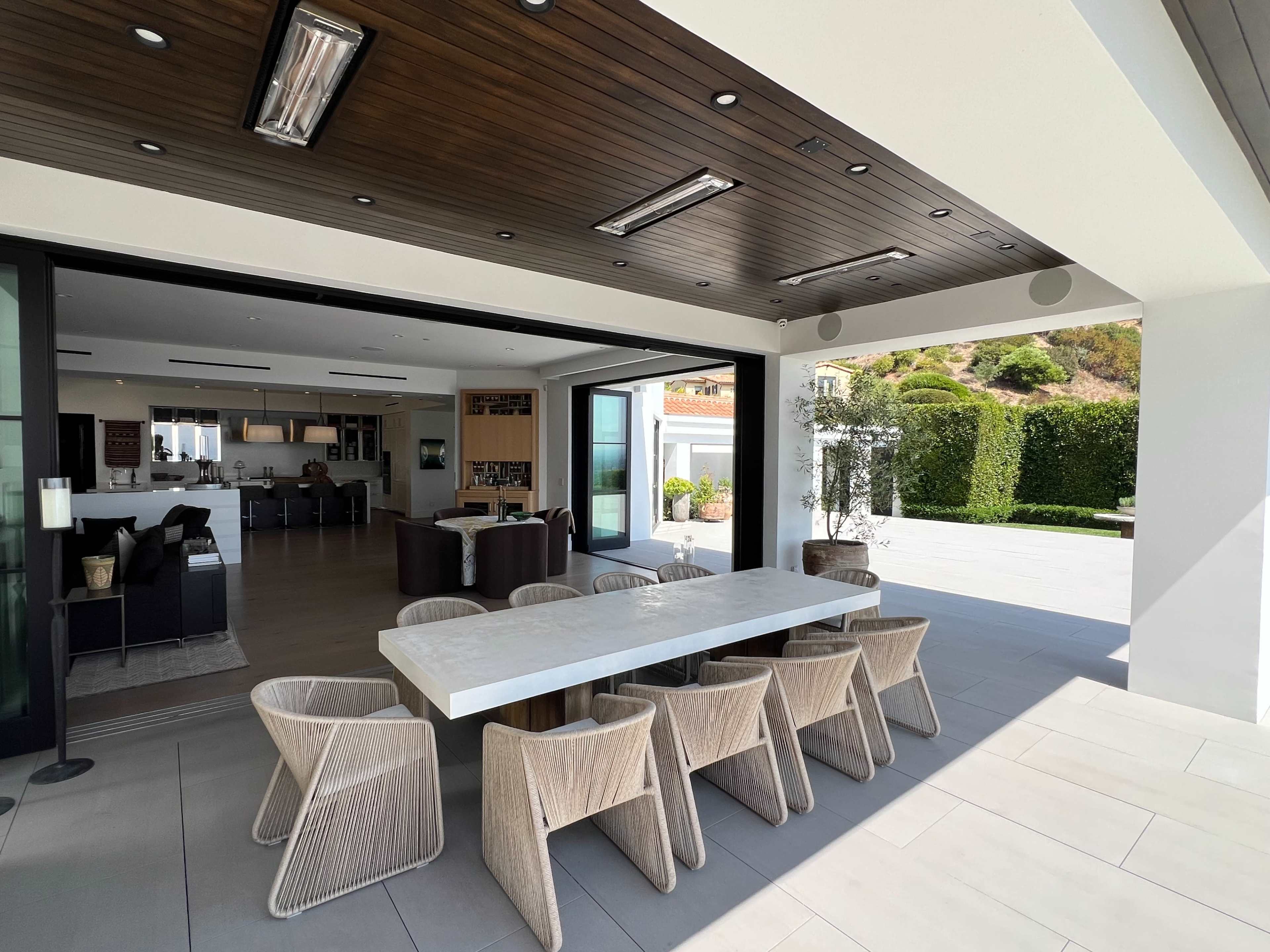 The image shows a spacious outdoor dining area with a white table surrounded by woven chairs, leading into a modern kitchen and living space.