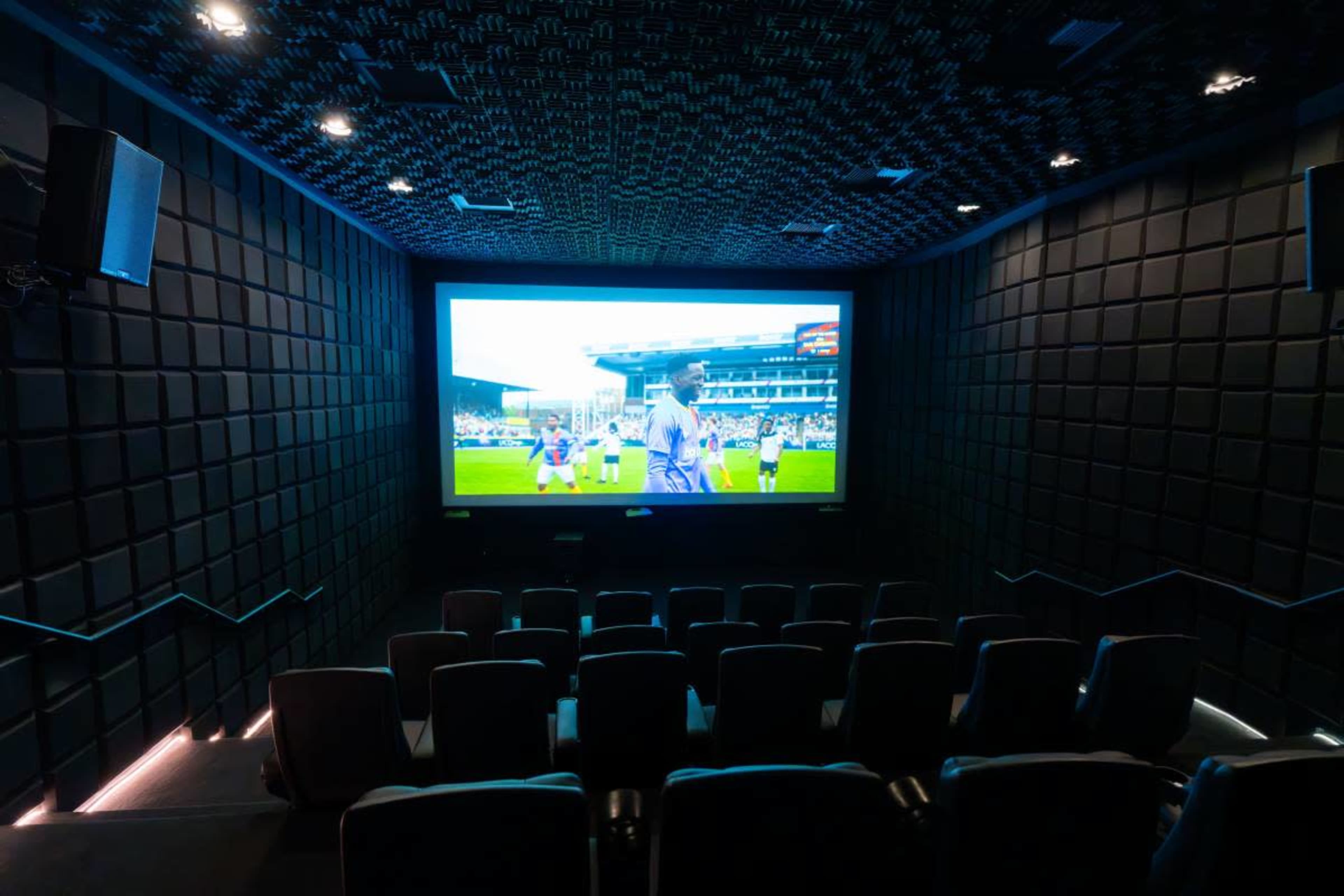 A small theater with tiered seating displays a large screen showing a live sports event.