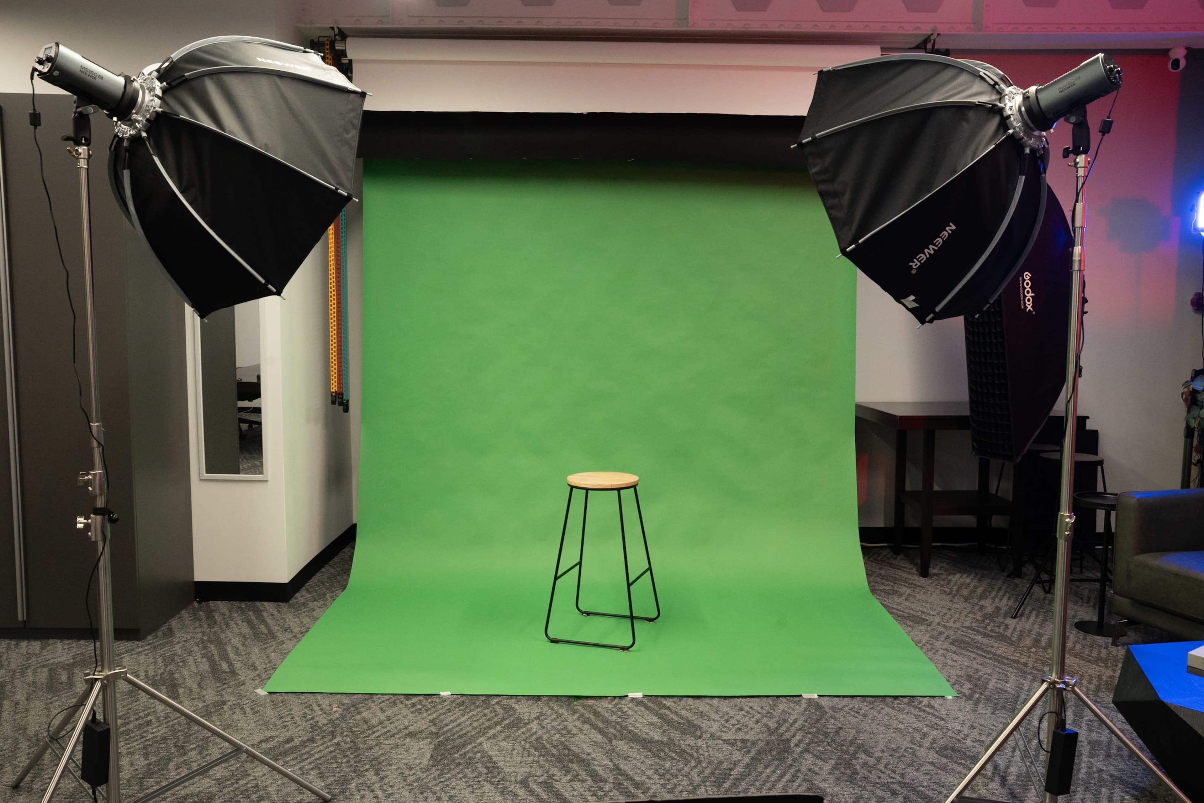 A photography studio is set up with two softbox lights and a green backdrop, featuring a single metal stool in front.