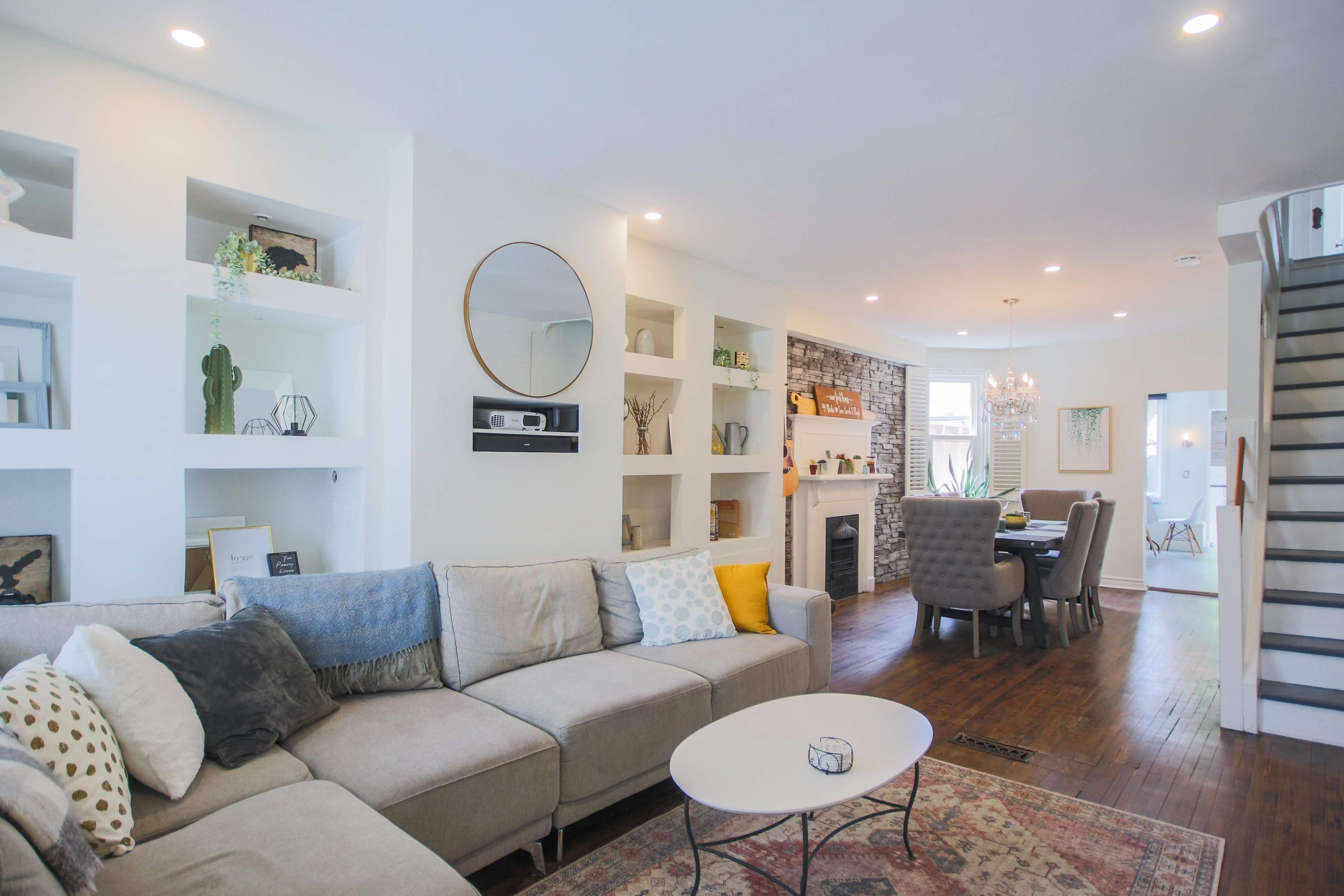 A modern living room with a gray sectional sofa, built-in shelves, a circular mirror, and a dining area in the background.