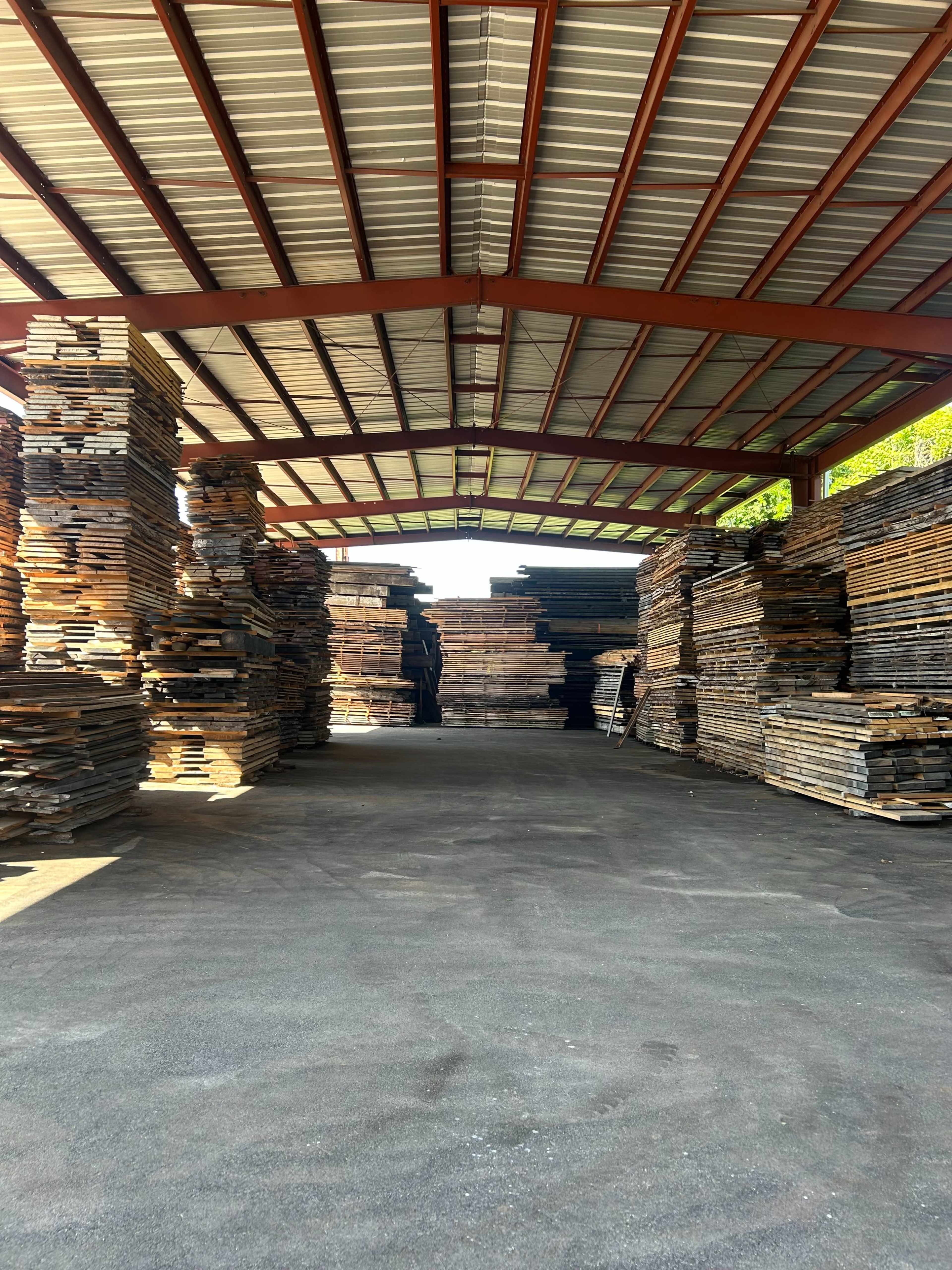 The image shows a covered warehouse with stacks of wooden planks neatly arranged on both sides.