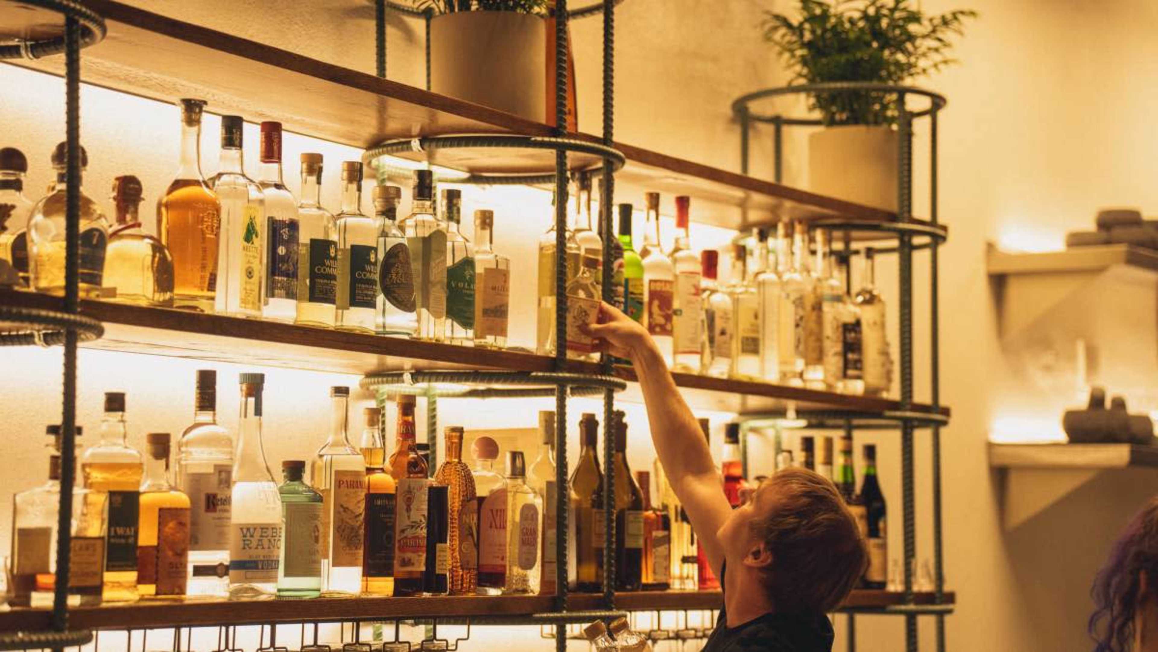 A person reaches for a bottle on a well-organized shelf filled with various liquor bottles.