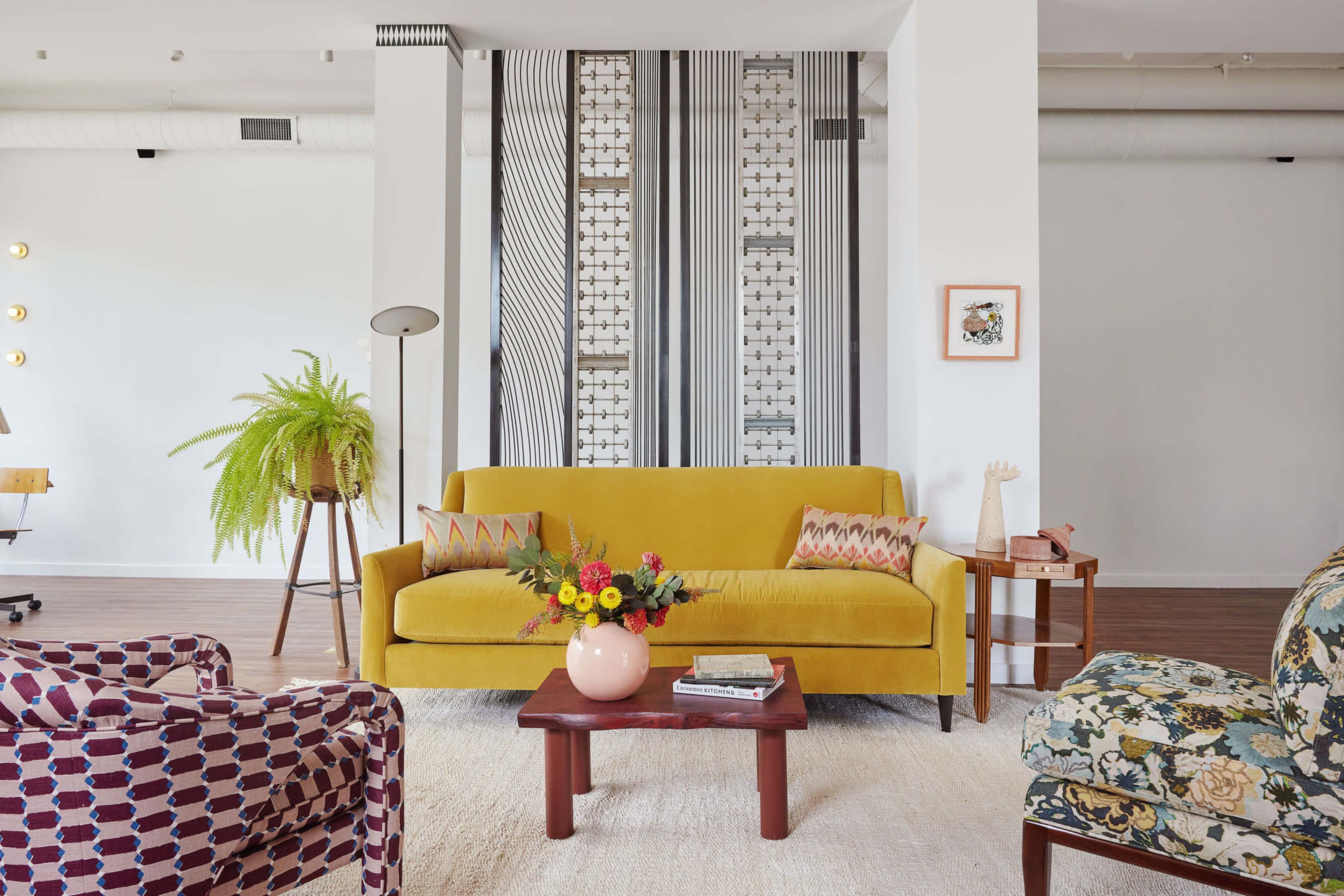 A vibrant living room features a yellow sofa, two distinct patterned chairs, a wooden coffee table with a flower arrangement, and a backdrop of striped wall decor.