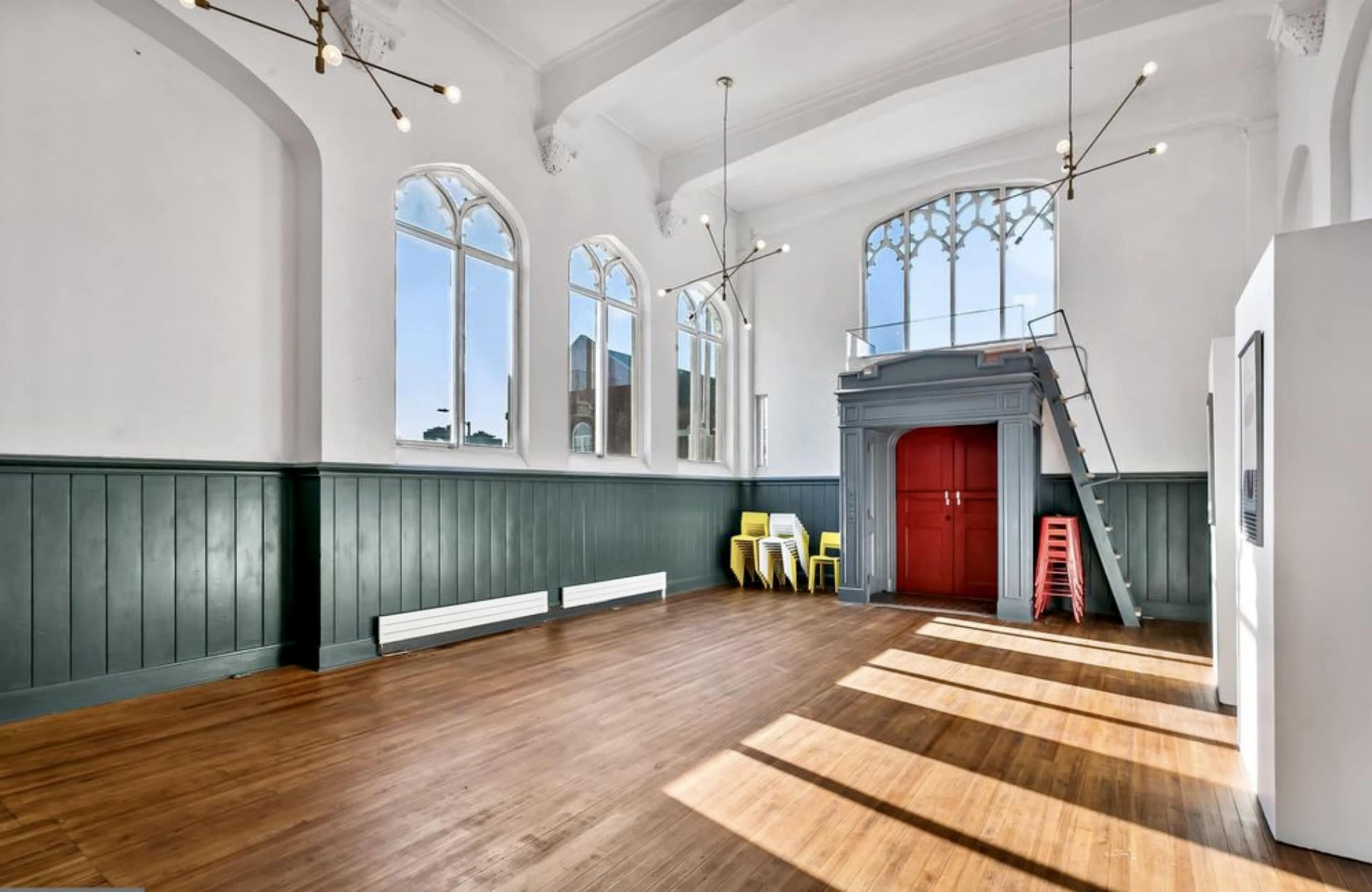 A large, bright interior space with tall windows, green paneling, wooden floors, and colorful chairs arranged near a red door and staircase.